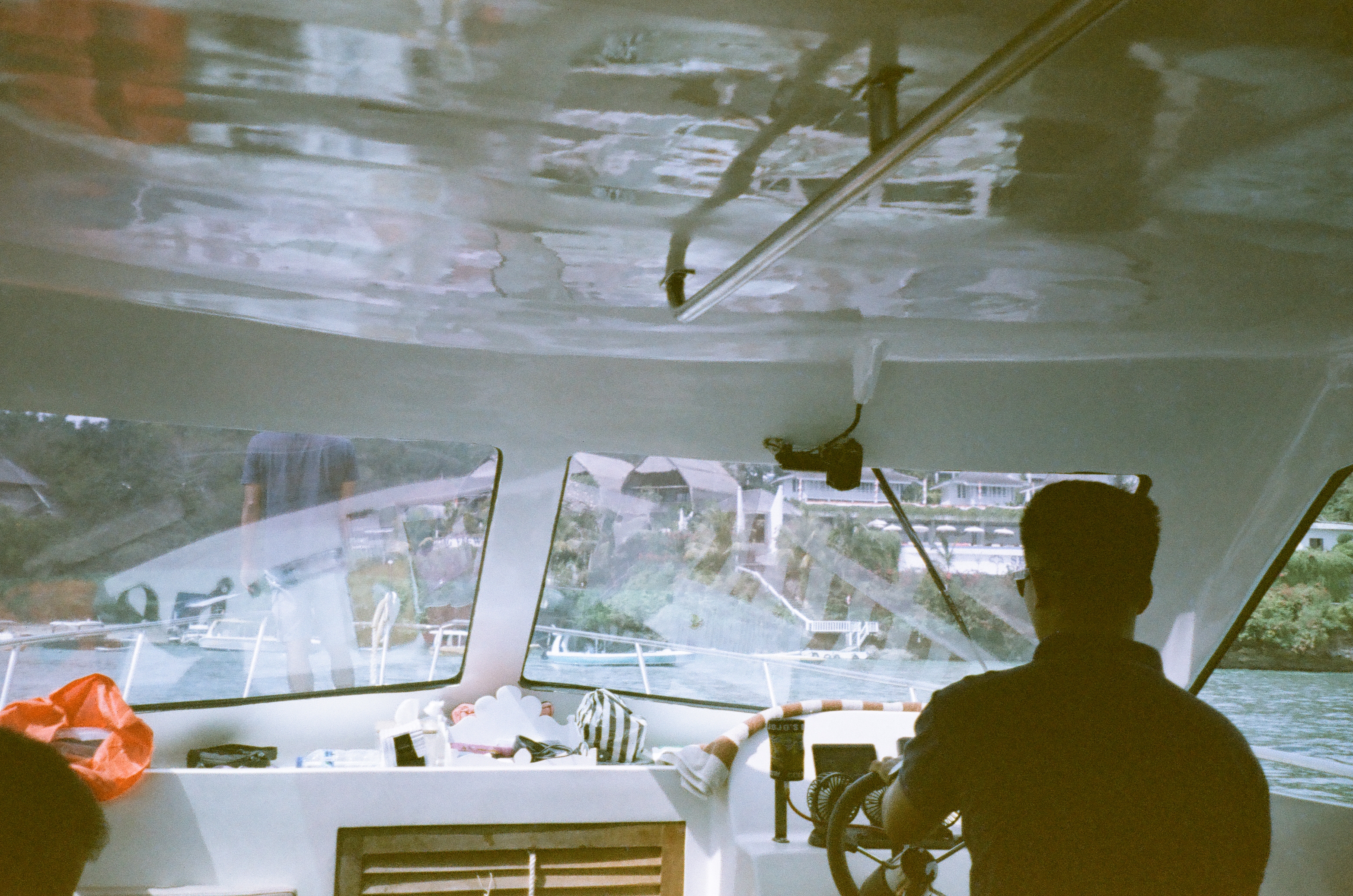 A person is at a boat's wheel while windows reveal water, boats, vegetation, and buildings along the shore.