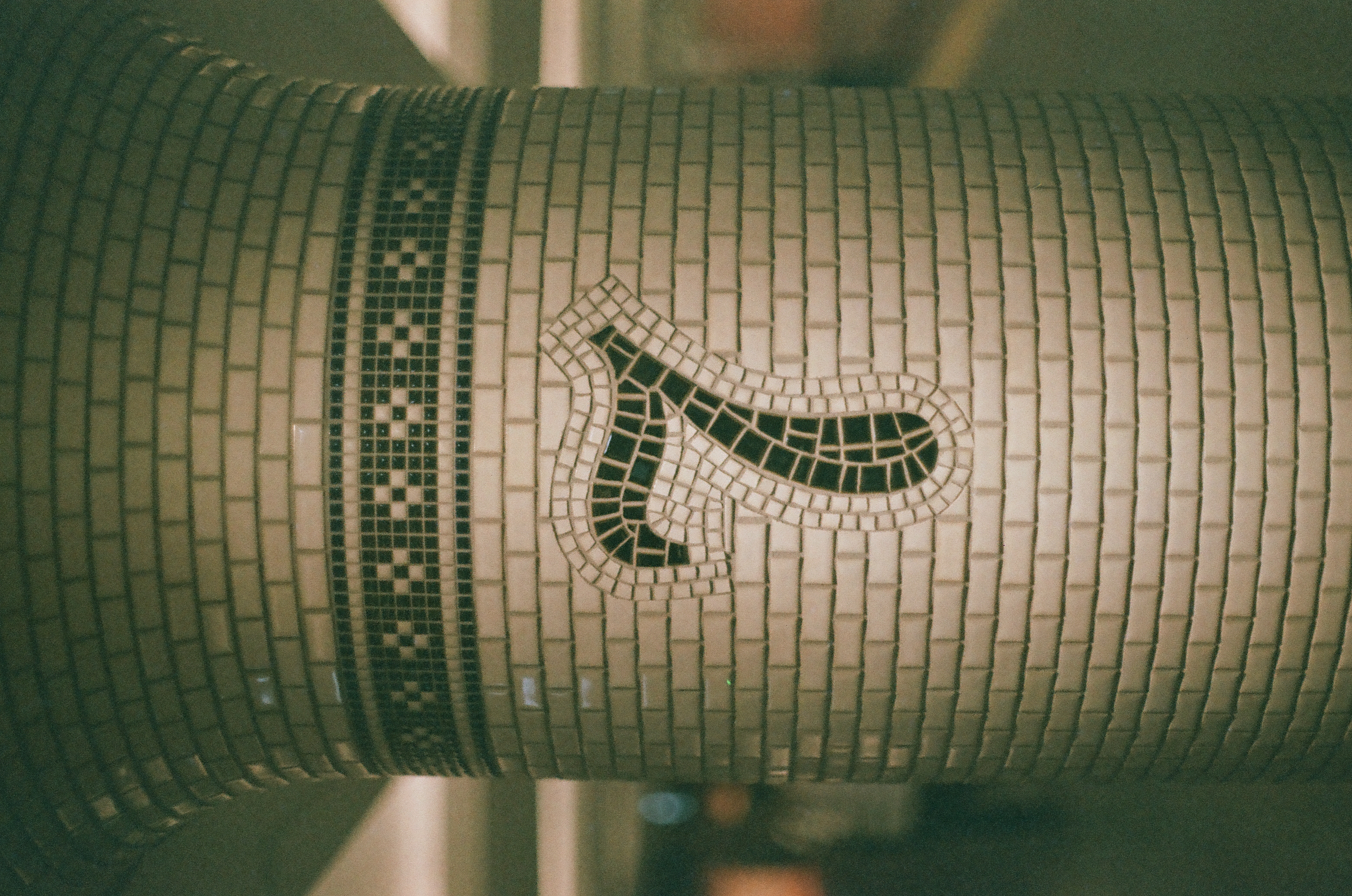 A tiled column shows the numeral seven formed from dark mosaic tesserae with a curled top stroke, beneath a checkered band, above a floor marked with a yellow line.