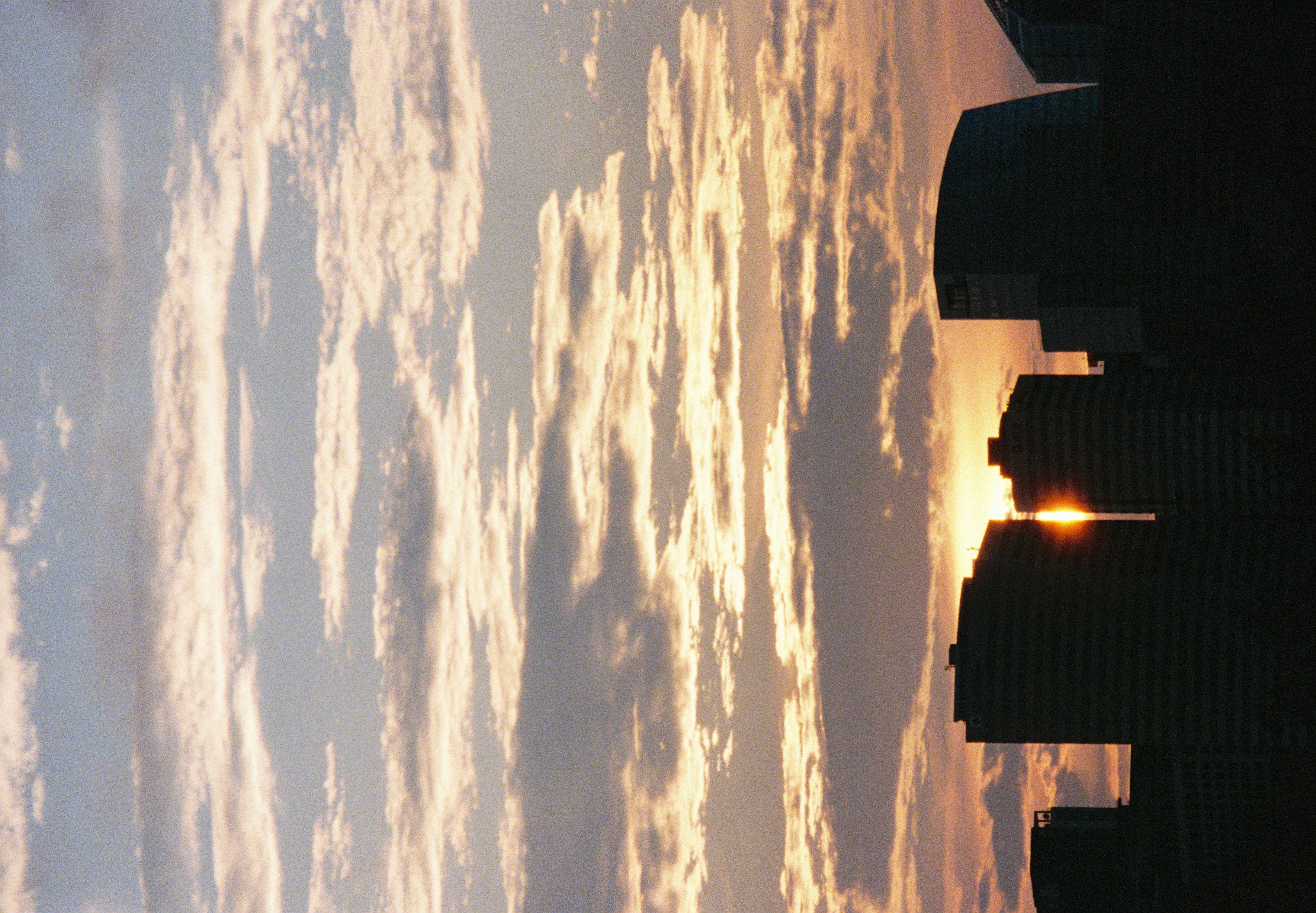 High-rise buildings silhouetted against a gold-and-gray cloud-banded sky, the low sun glowing in the gap between two towers beneath rows of small scattered clouds.