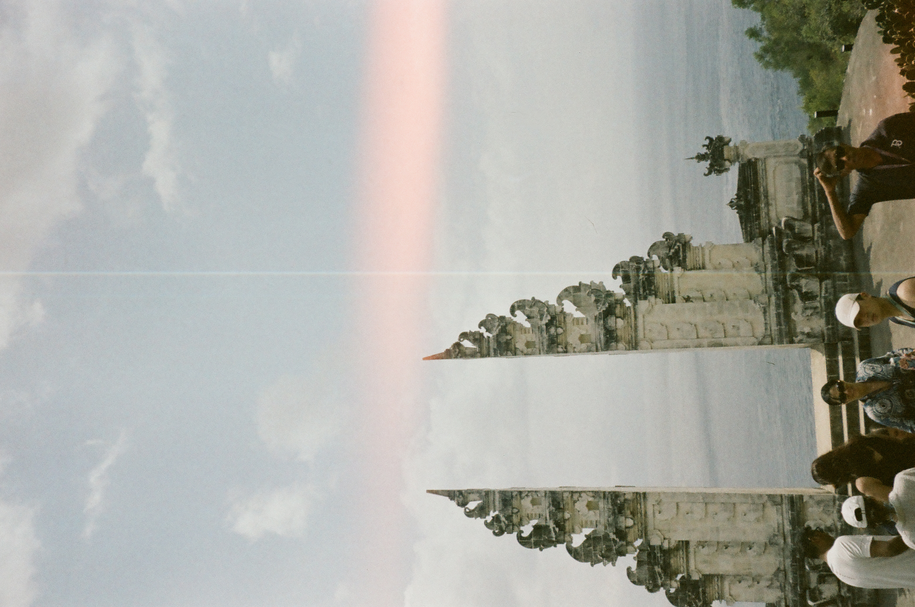 Several visitors stand below an ornate split stone gateway as cloudy sky and a pale vertical line fill the frame.