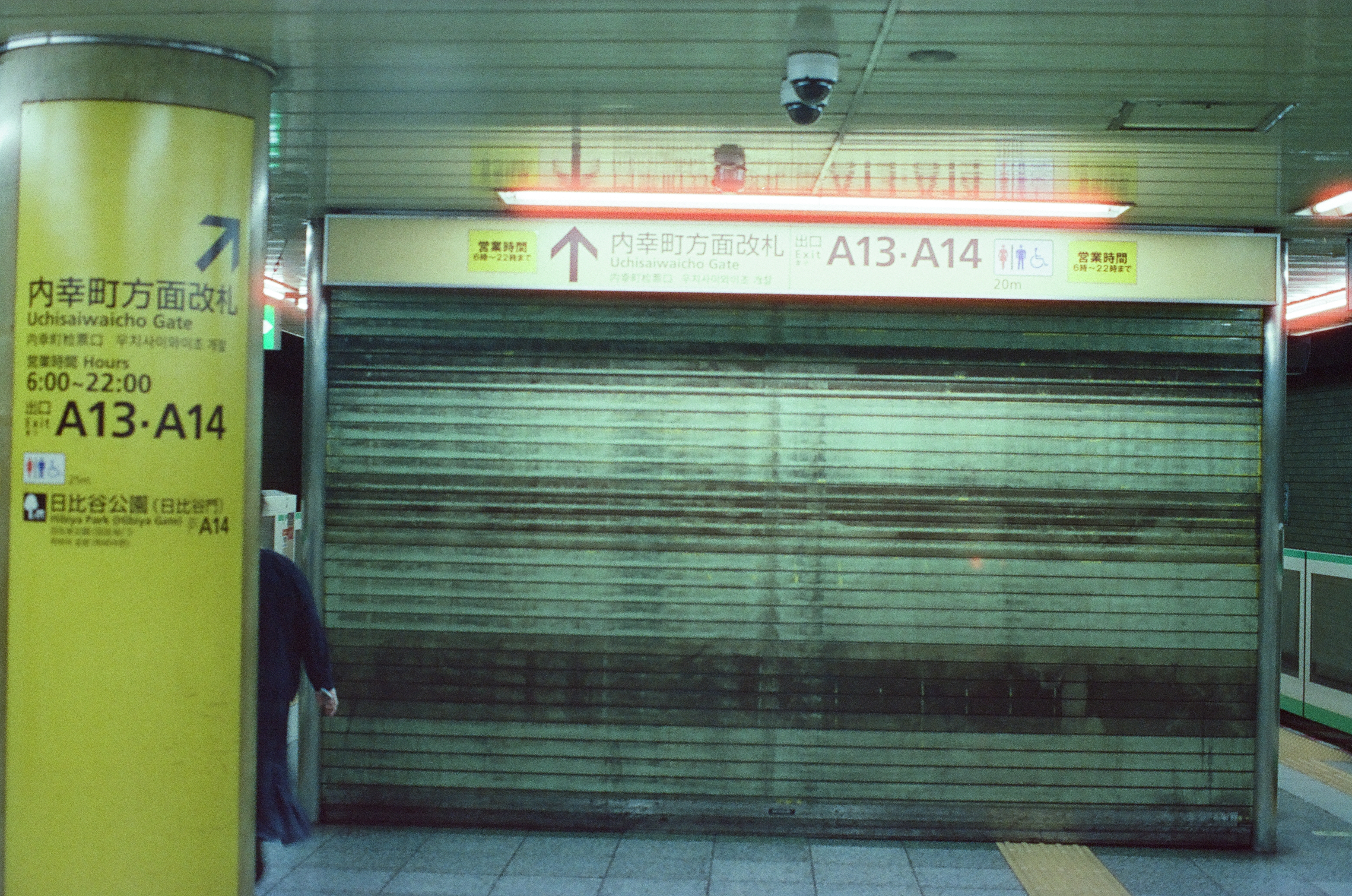 A corrugated metal shutter seals a subway ticket gate beneath yellow signage reading Uchisaiwaicho Gate, Exit A13 and A14, as a security camera watches from the ceiling.