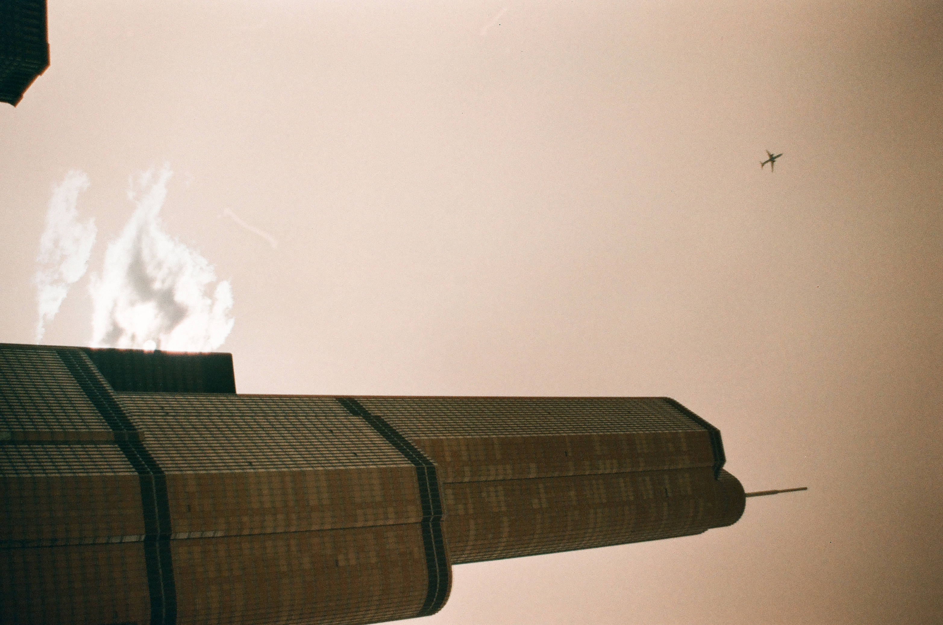 A boxy high-rise narrows against a hazy pink sky, a faint contrail cloud to one side and a small airplane crossing near the frame's edge.