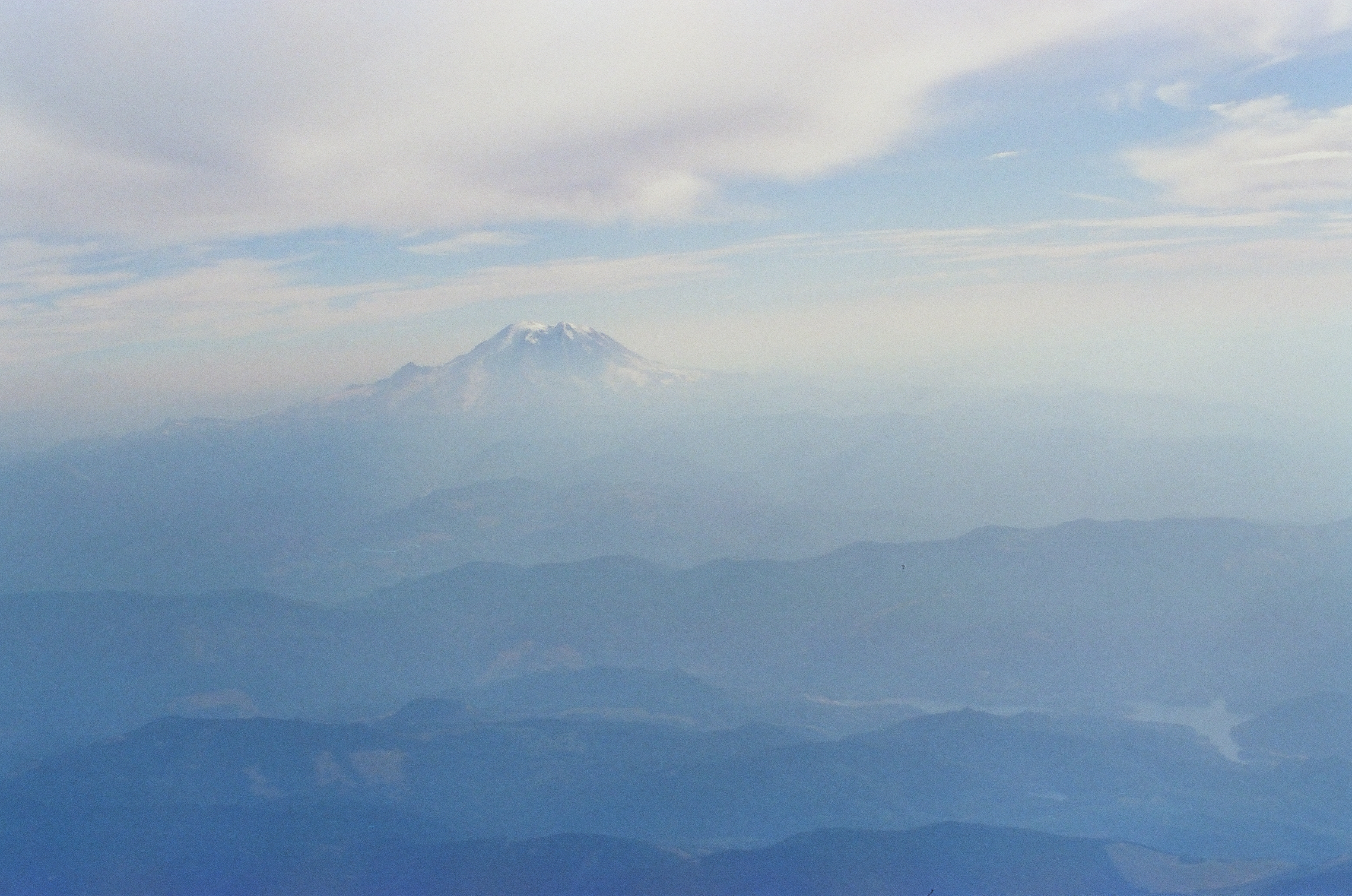 A snow-capped peak rises beside a smaller, lower ridge above layered blue mountains, with a faint pale patch visible in a valley below.