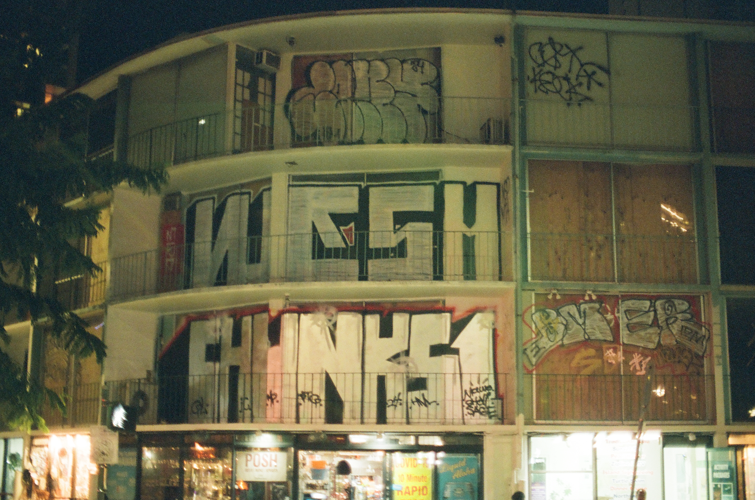Large graffiti lettering covers the balconies and walls of a dark vacant building above bright shops.