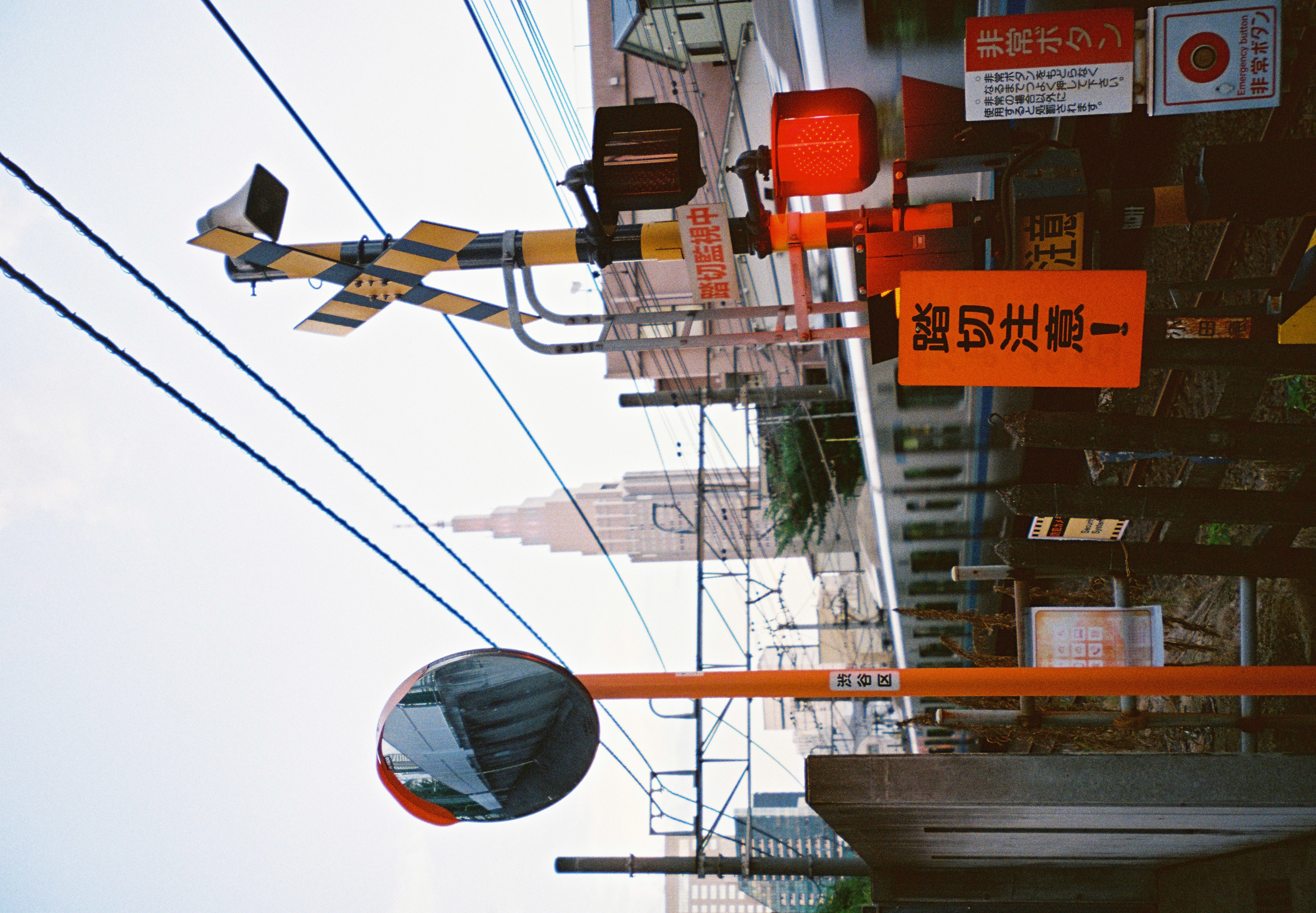 A convex mirror on an orange pole, a striped crossbuck, a red lamp, and an orange sign reading 踏切注意 stand before a blurred train and a distant tower.