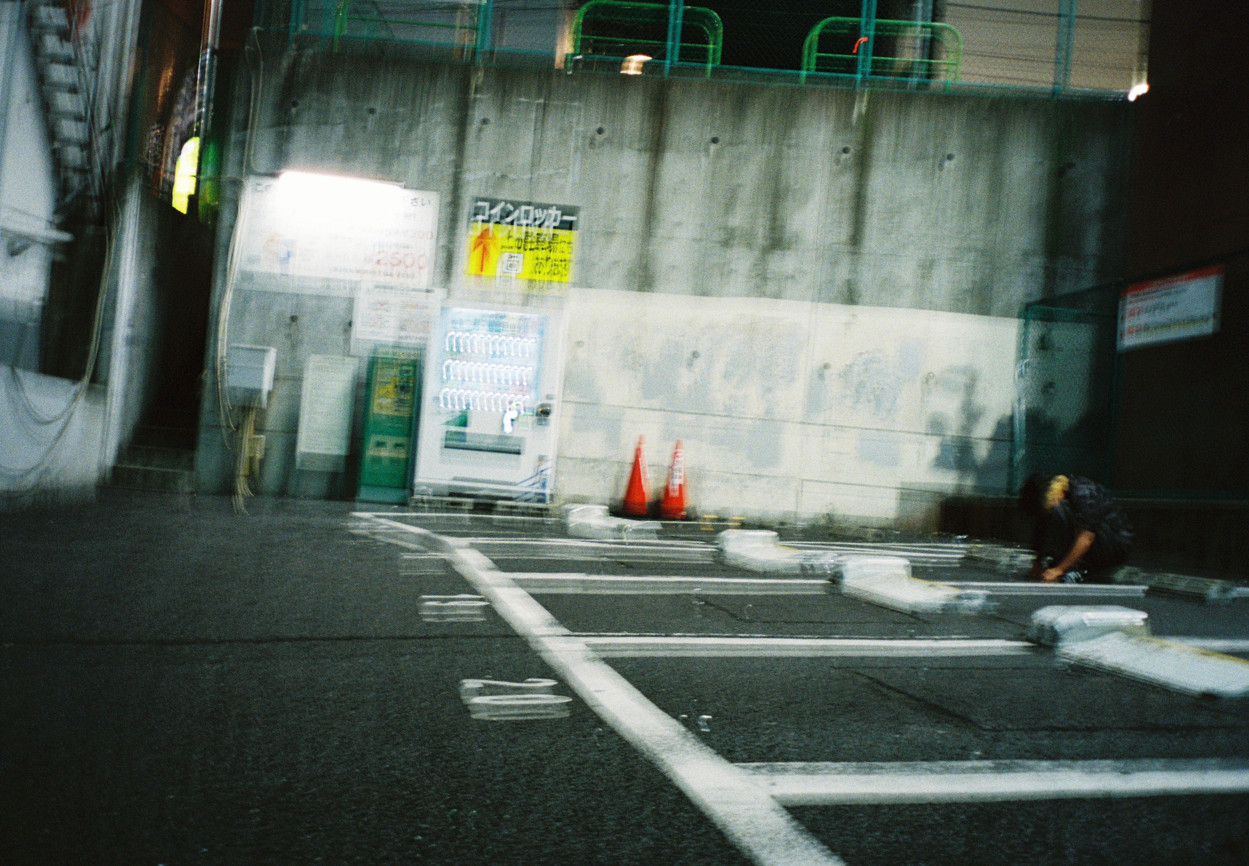 An empty nighttime parking lot holds a lit vending machine, illuminated signage, two orange traffic cones, and a crouching figure at right.