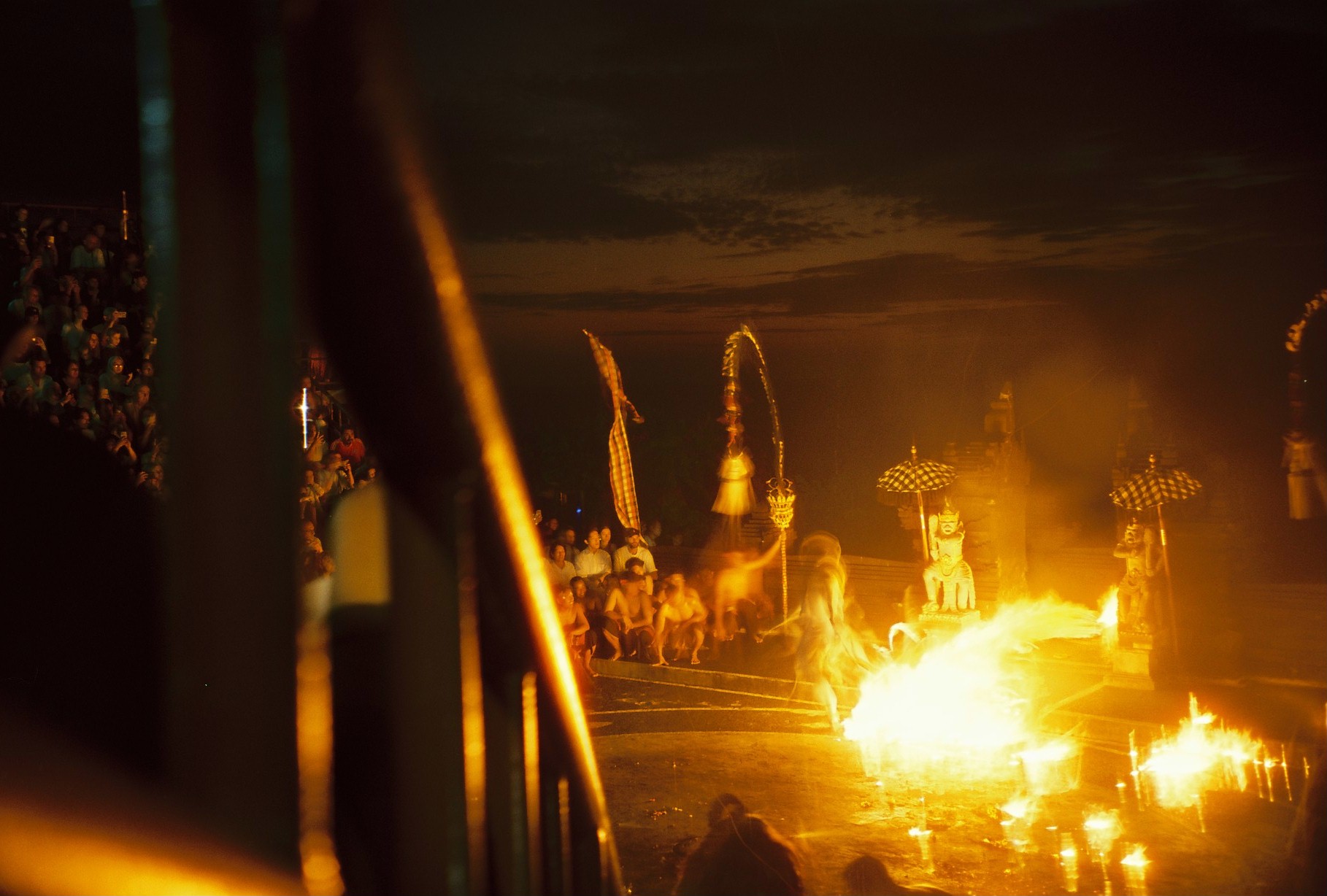 Several orange fires burn across a dark central area while seated spectators and tall decorative forms line the background.
