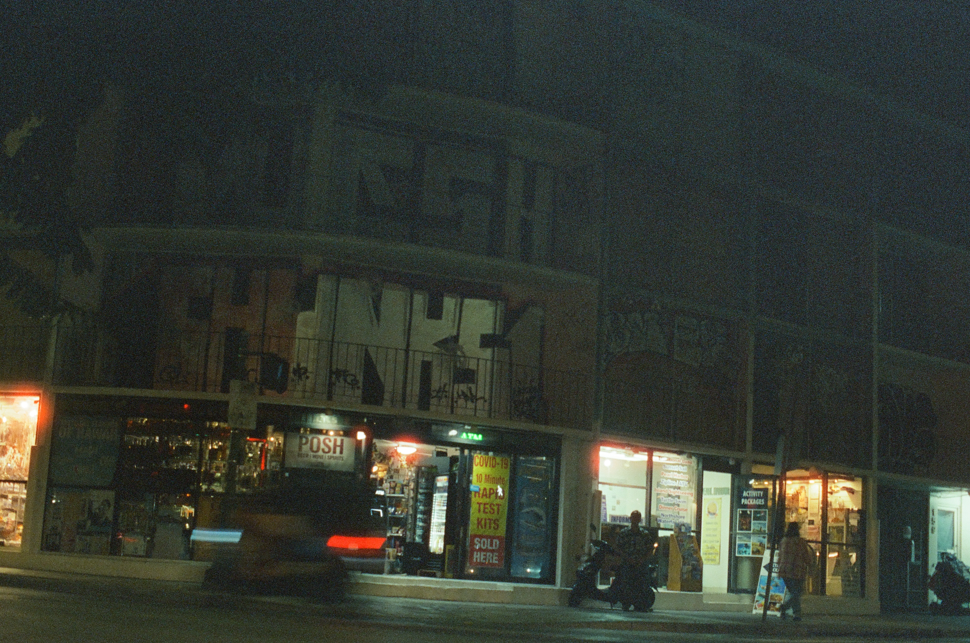 A row of brightly lit shops faces a dark road as a vehicle passes through the center.