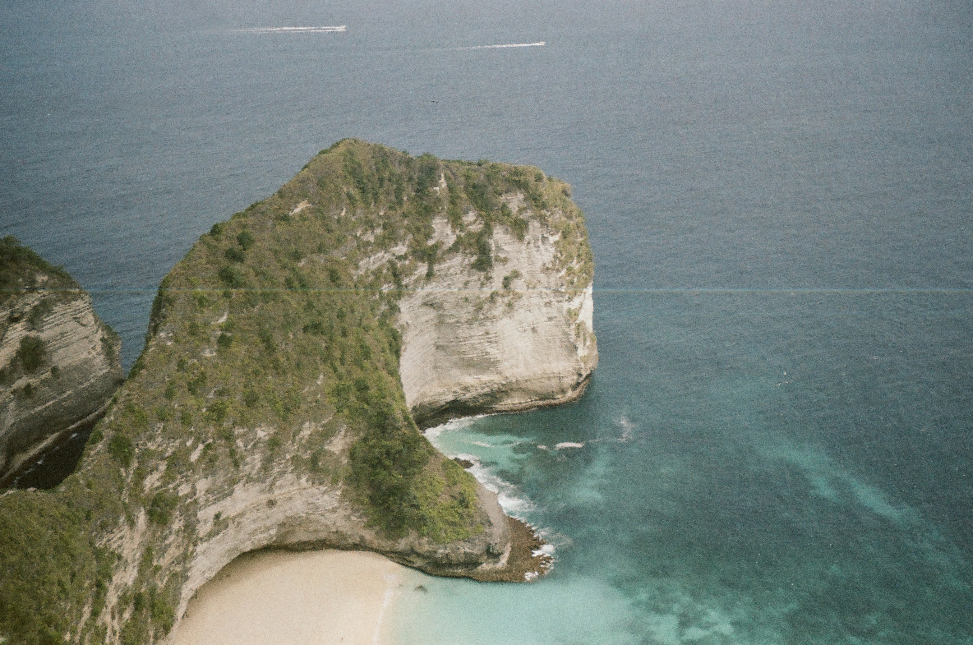 A broad green and pale rock headland curves around a sandy cove amid blue and turquoise water.