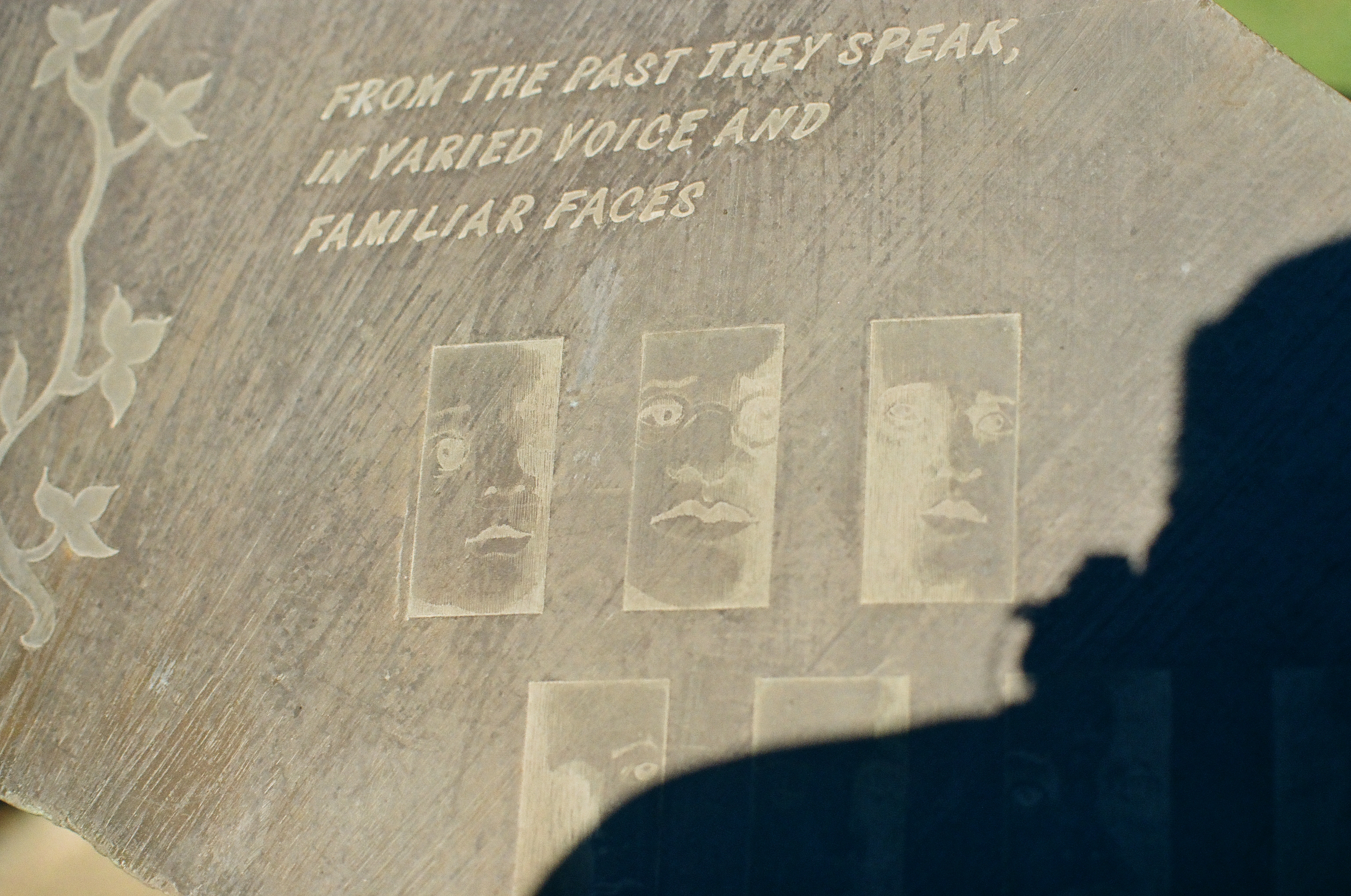 An engraved stone slab reads FROM THE PAST THEY SPEAK, IN VARIED VOICE AND FAMILIAR FACES above small panels etched with faces.