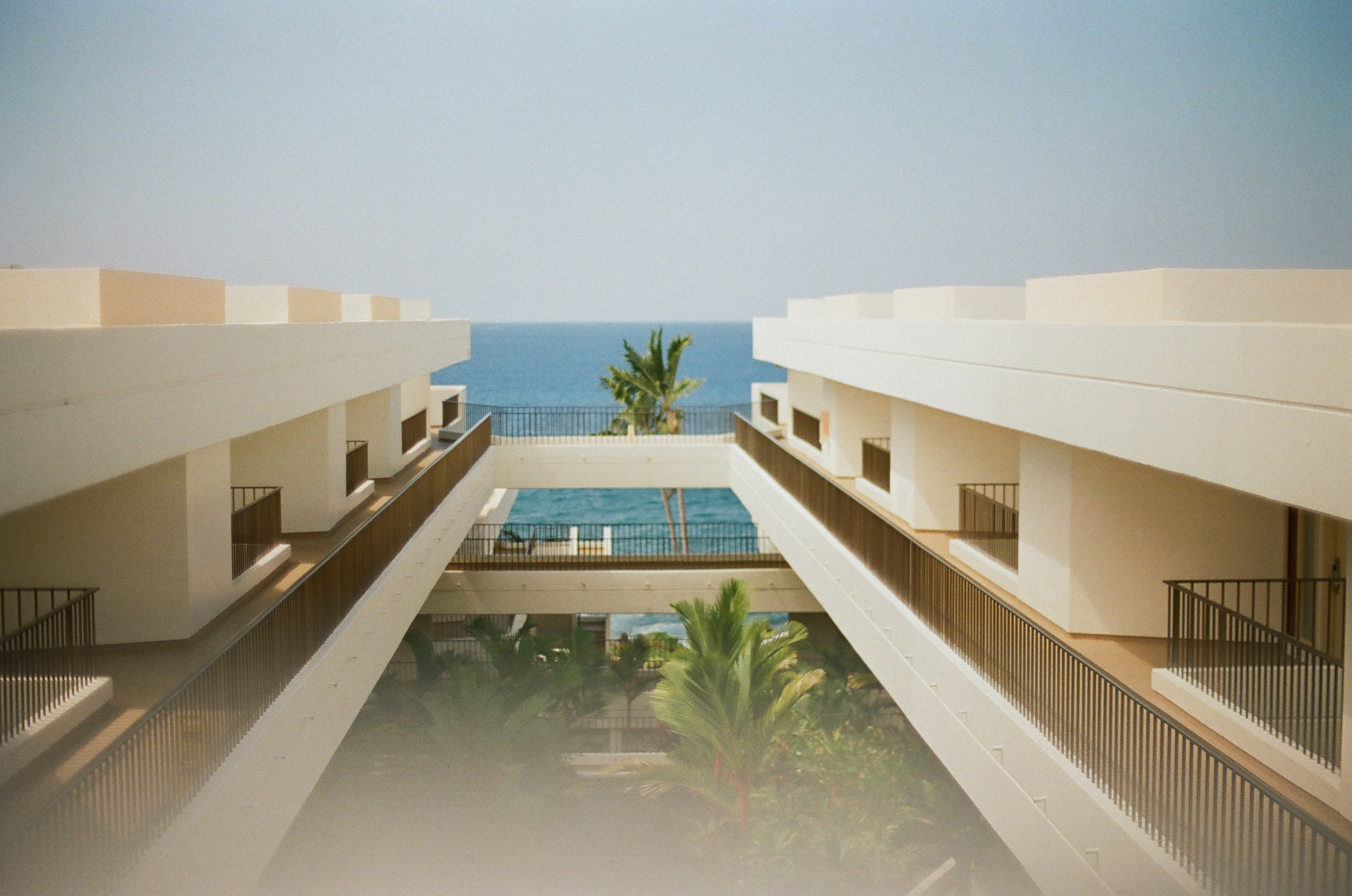Two pale building wings and dark balcony rails converge toward palm trees, blue water, and open sky.