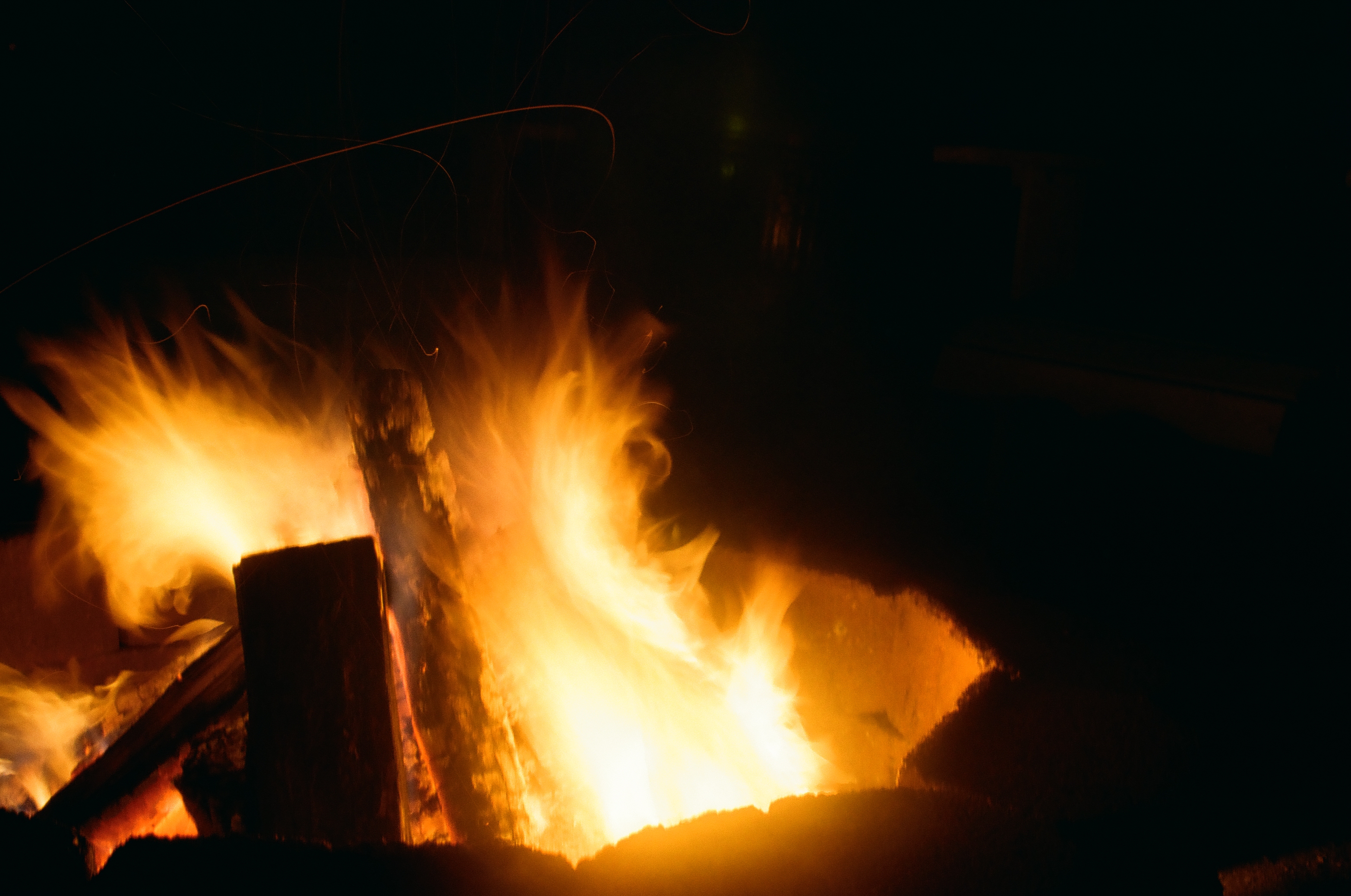 Thin curling sparks rise above a fire built over crossed logs, with a dark angled shape visible at the upper right.