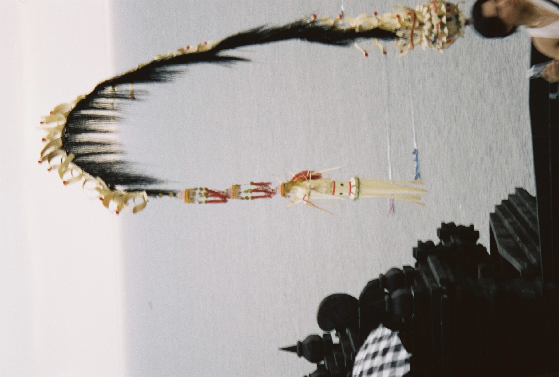 A curved ceremonial ornament trimmed with black fiber fringe and beaded tassels hangs against a pale sky and grey sea beside a dark carved temple roof.
