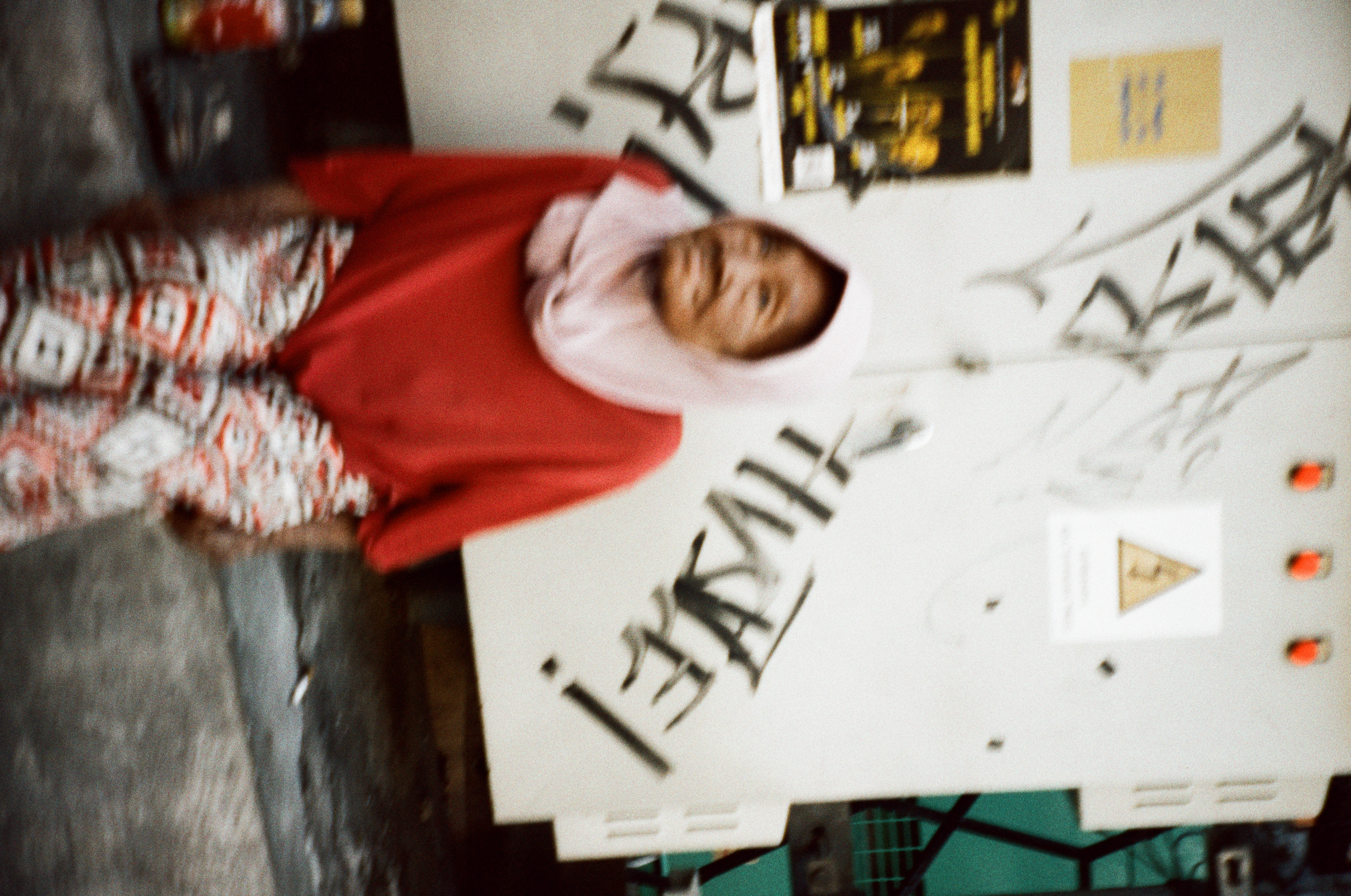 An elderly woman in a pale headscarf, red top, and patterned sarong rests in front of a white cabinet covered in dark graffiti tags.