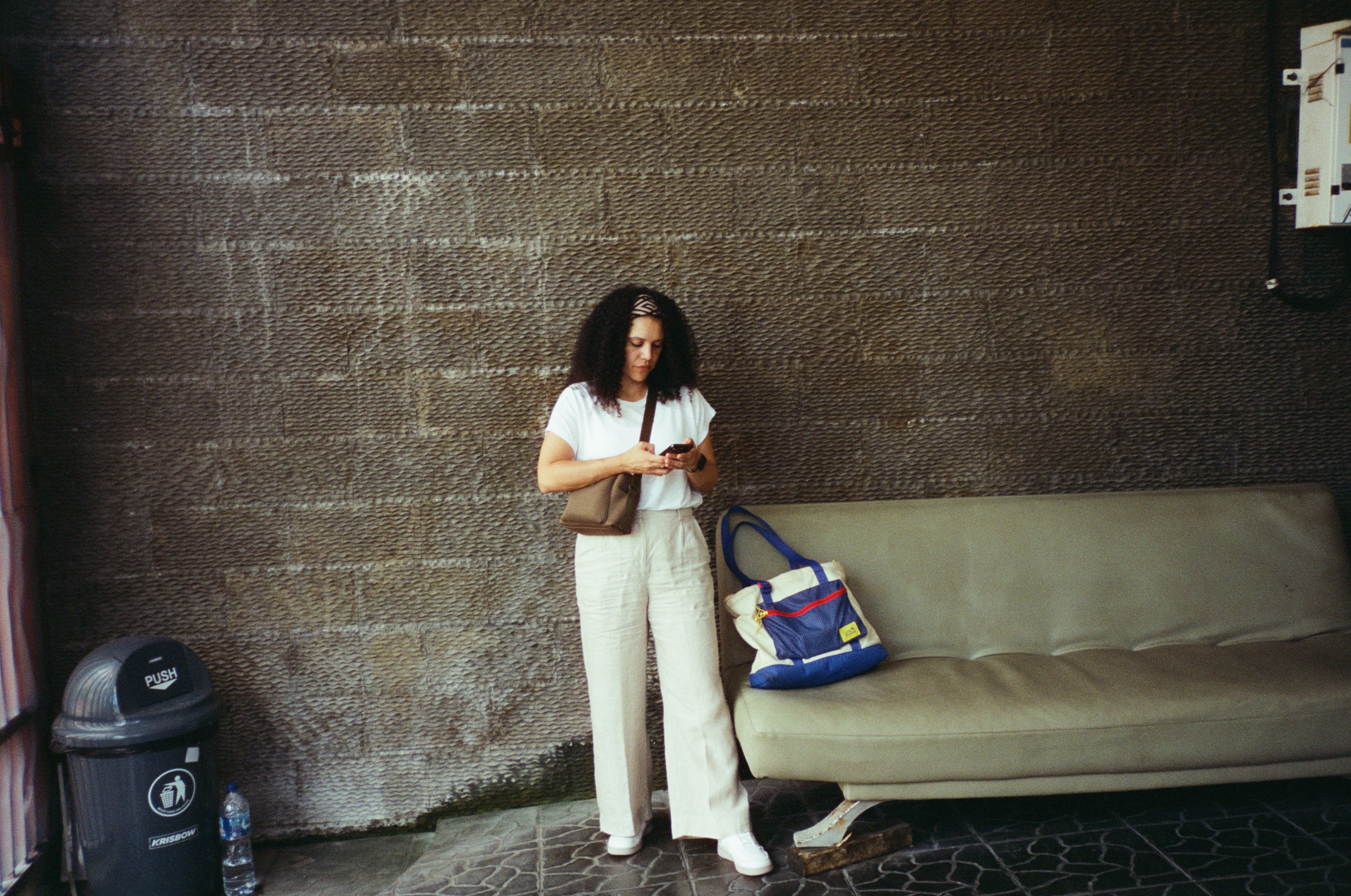 A woman with curly hair and a patterned headband looks at her phone beside a pale green couch holding a blue and cream tote bag.