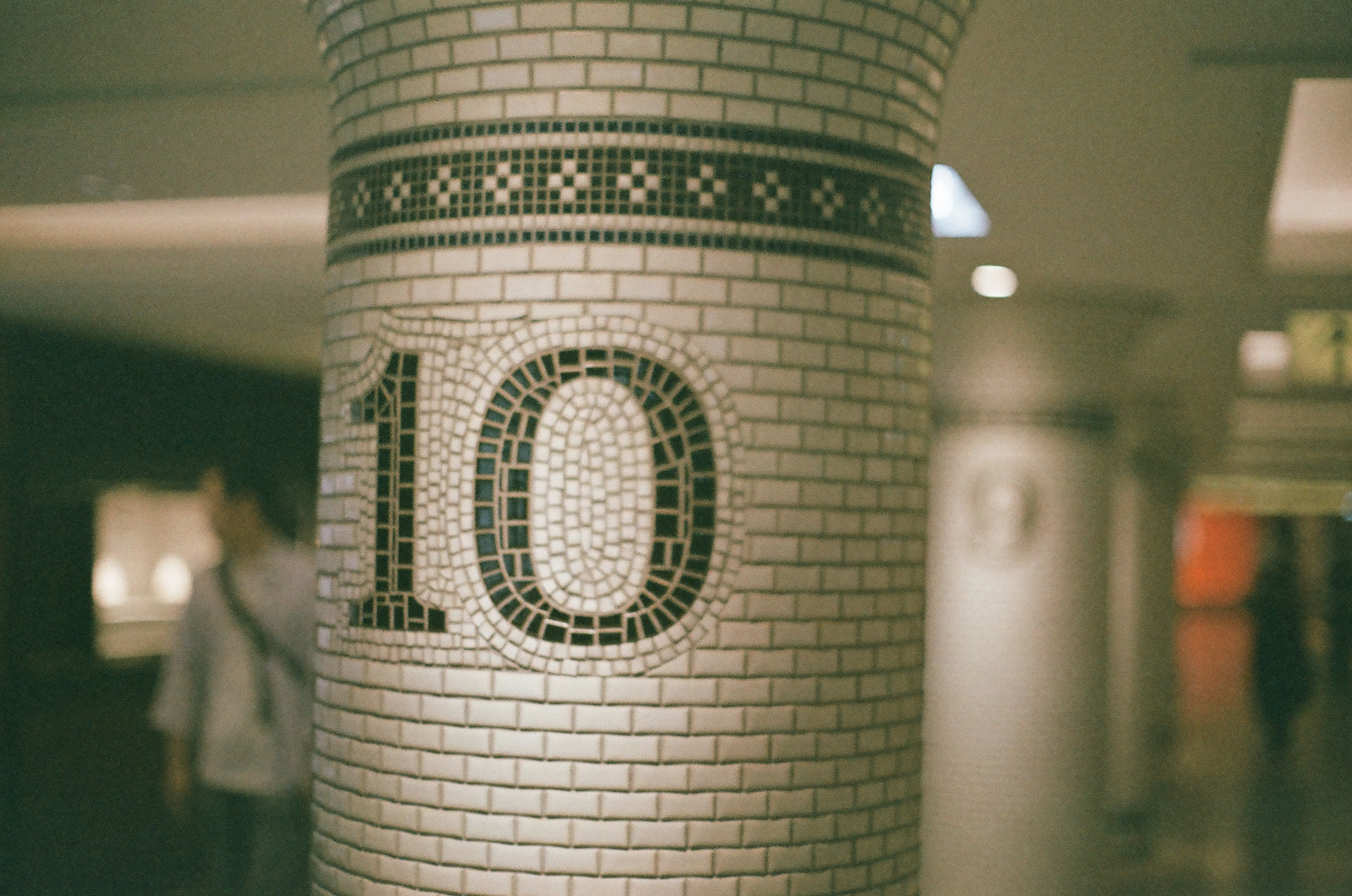 A round column covered in white brick-pattern tile displays the numeral ten in dark mosaic tesserae beneath a checkered band, with a blurred figure passing behind it.