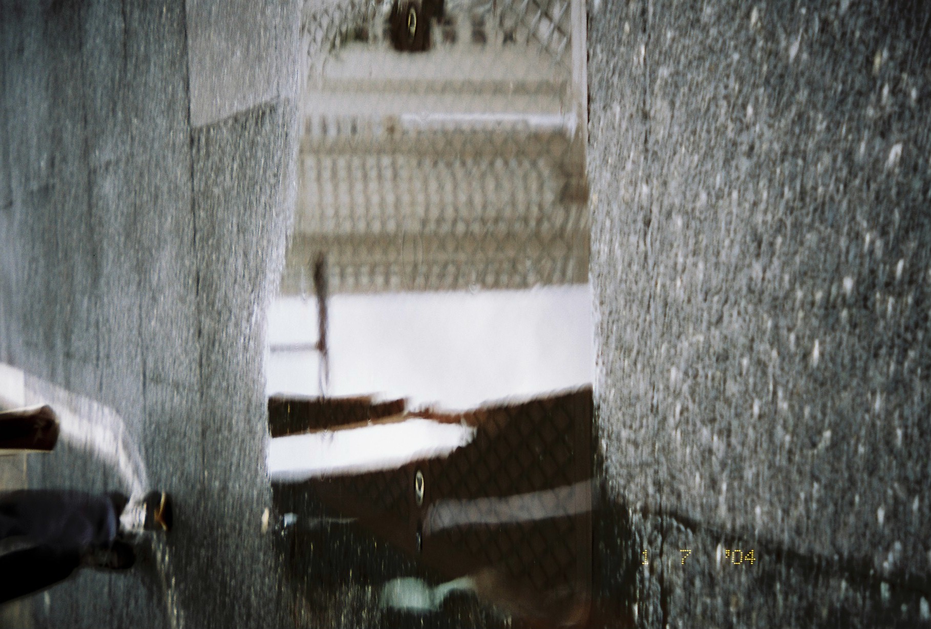 A band of standing water on dark asphalt reflects a gridded facade and pale sky, with inverted legs and shoes of passing pedestrians at the lower right.