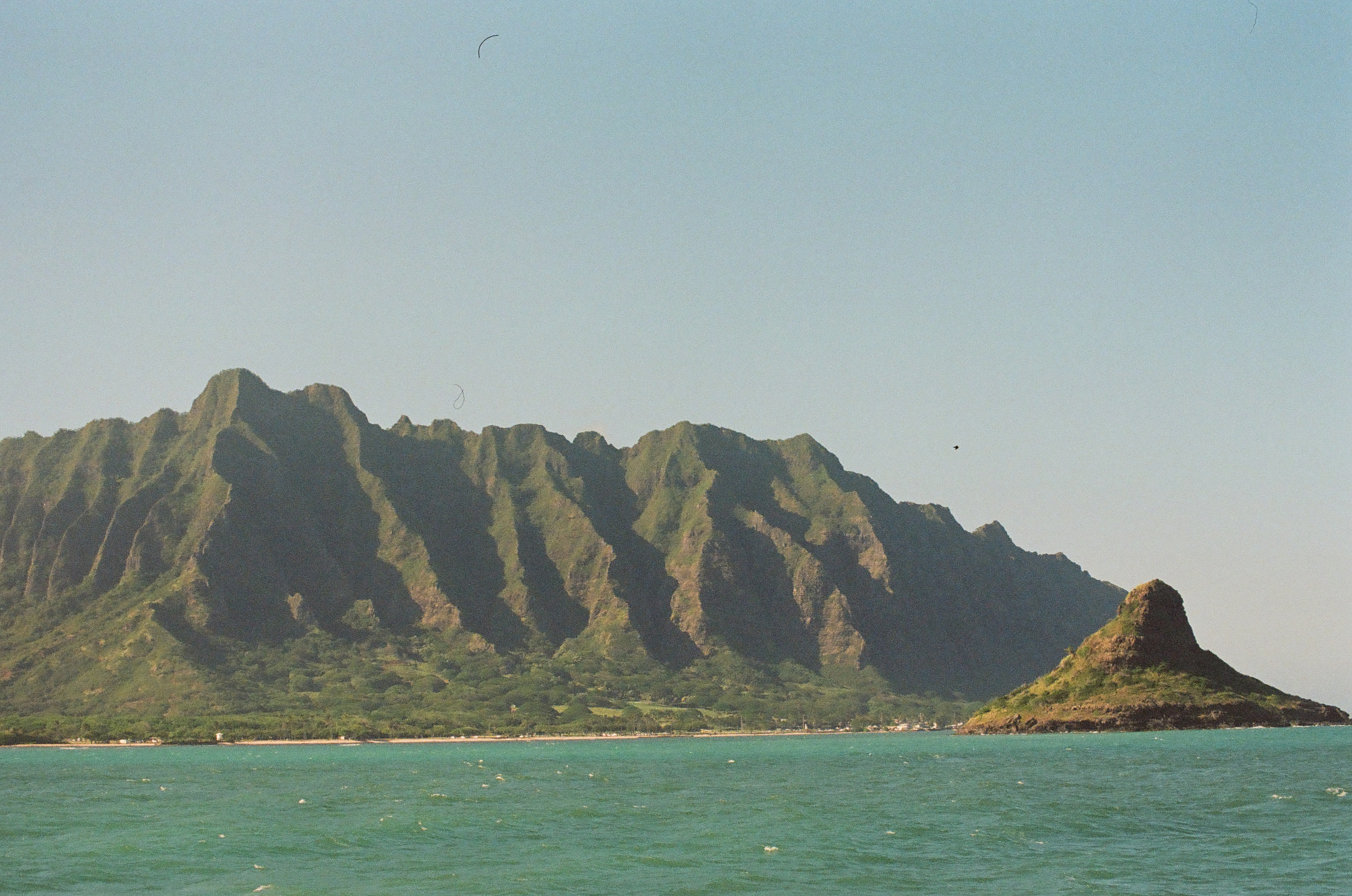 A long ridged green mountain range meets a rounded rocky peninsula on the right, with turquoise water and a hazy pale sky above.