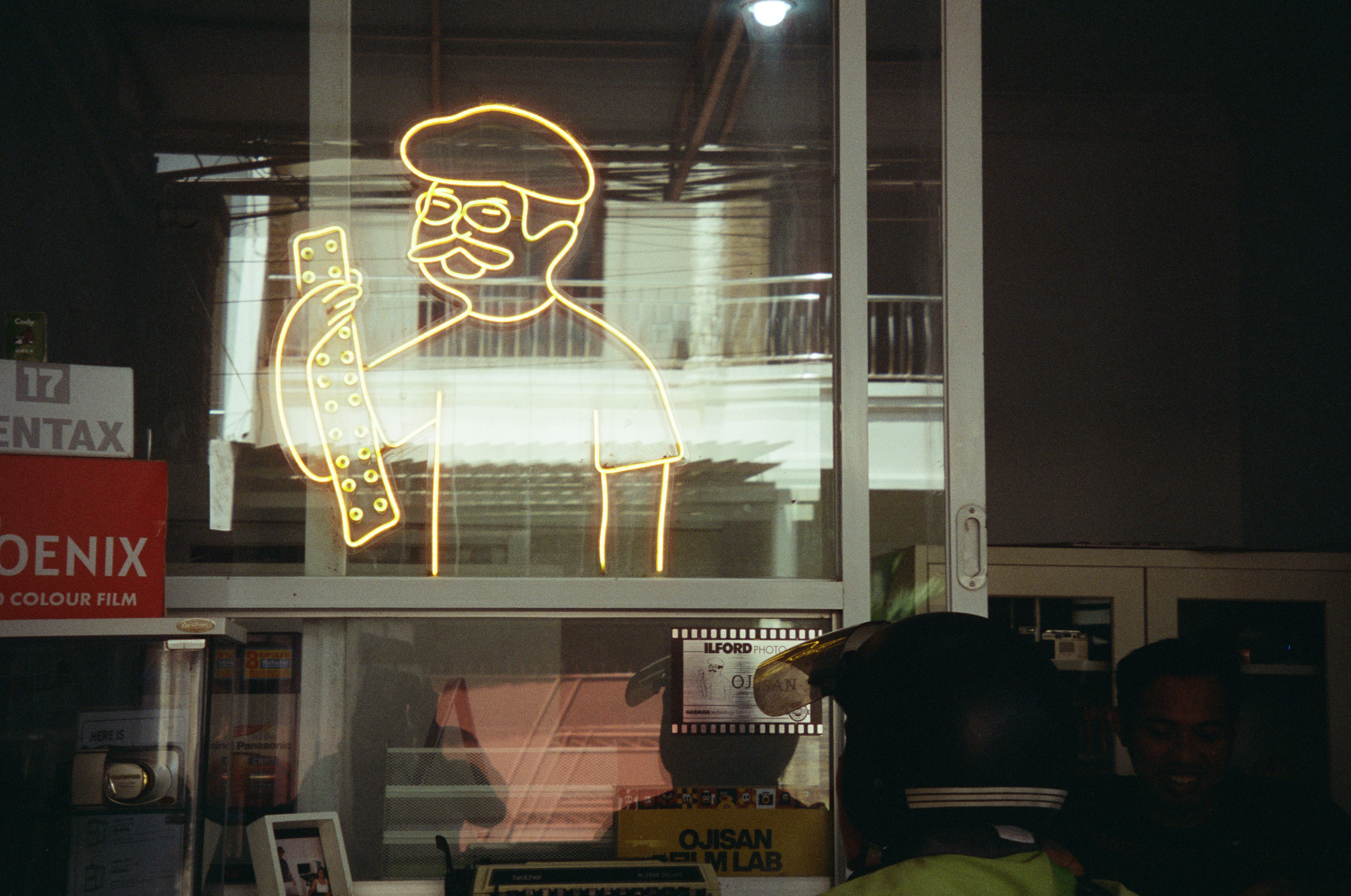 Yellow neon outline of a mustachioed man in a beret and glasses holding a dotted film strip glows in a window above shelves, a helmeted head, and a smiling face.