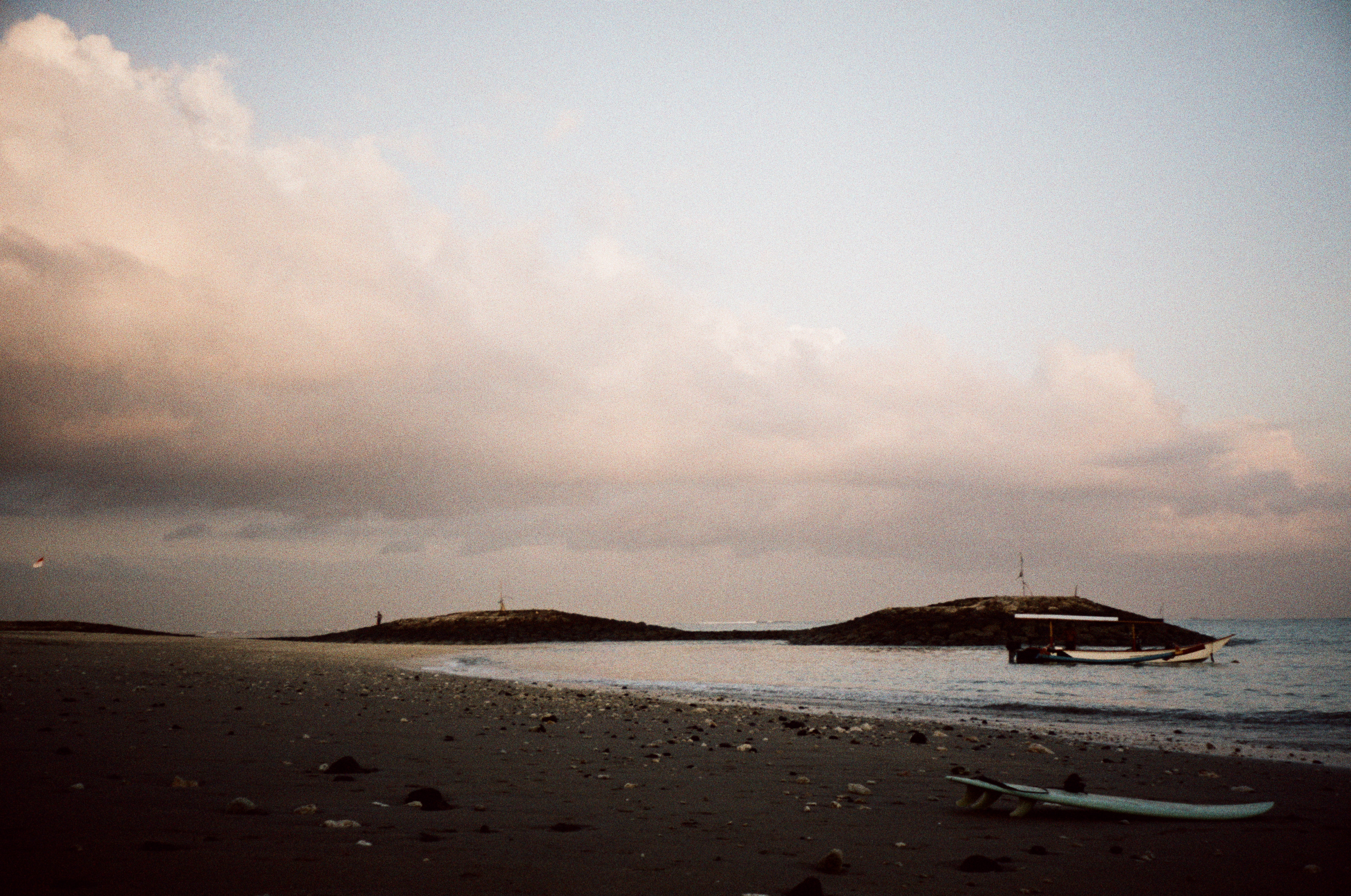 Dark sand scattered with shells and stones slopes toward calm water, with a pale surfboard in the foreground and a canopied boat near rocks.