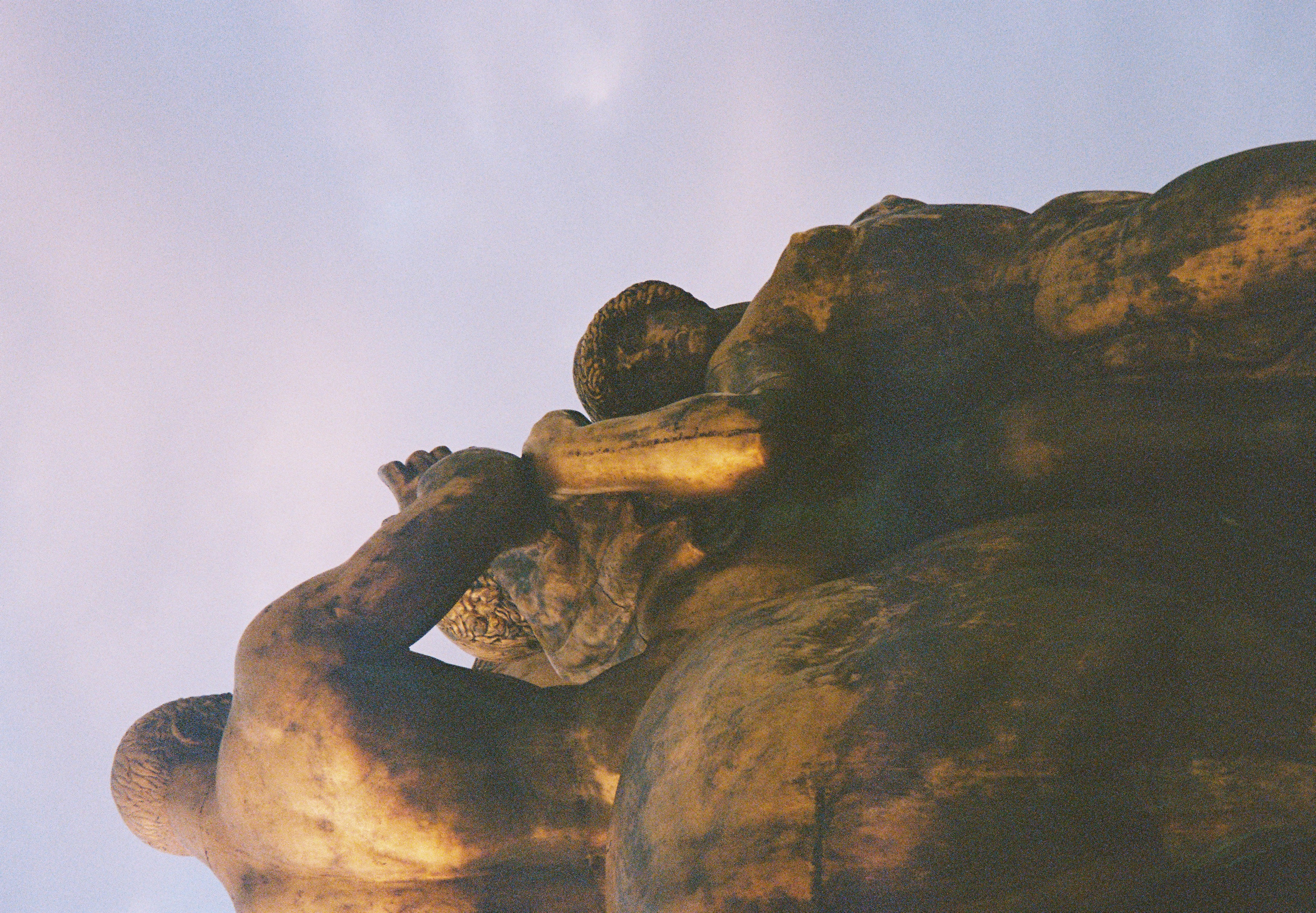 A bronze-toned group of muscular sculpted figures rises against a softly colored evening sky.