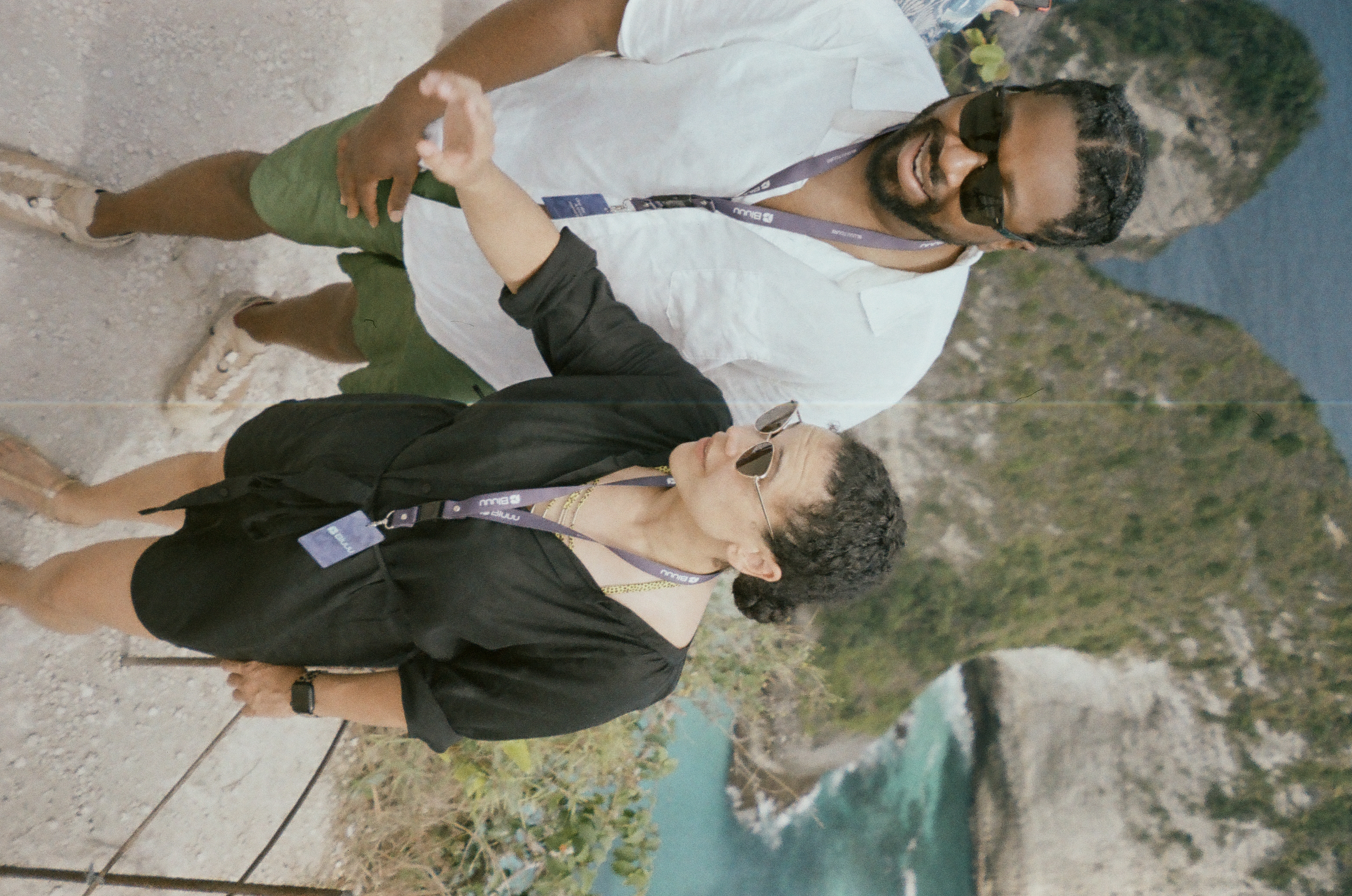 Two people climb a steep path above a turquoise cove bordered by a rugged green cliff.