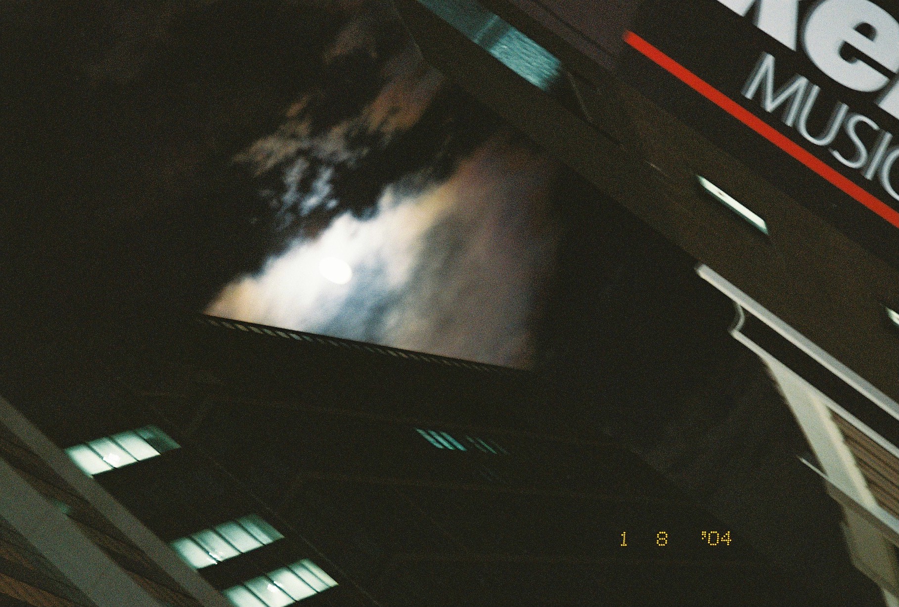 The moon glows behind moving clouds beside a dark building edge, with a partial sign reading Music cropped at the frame's top right.