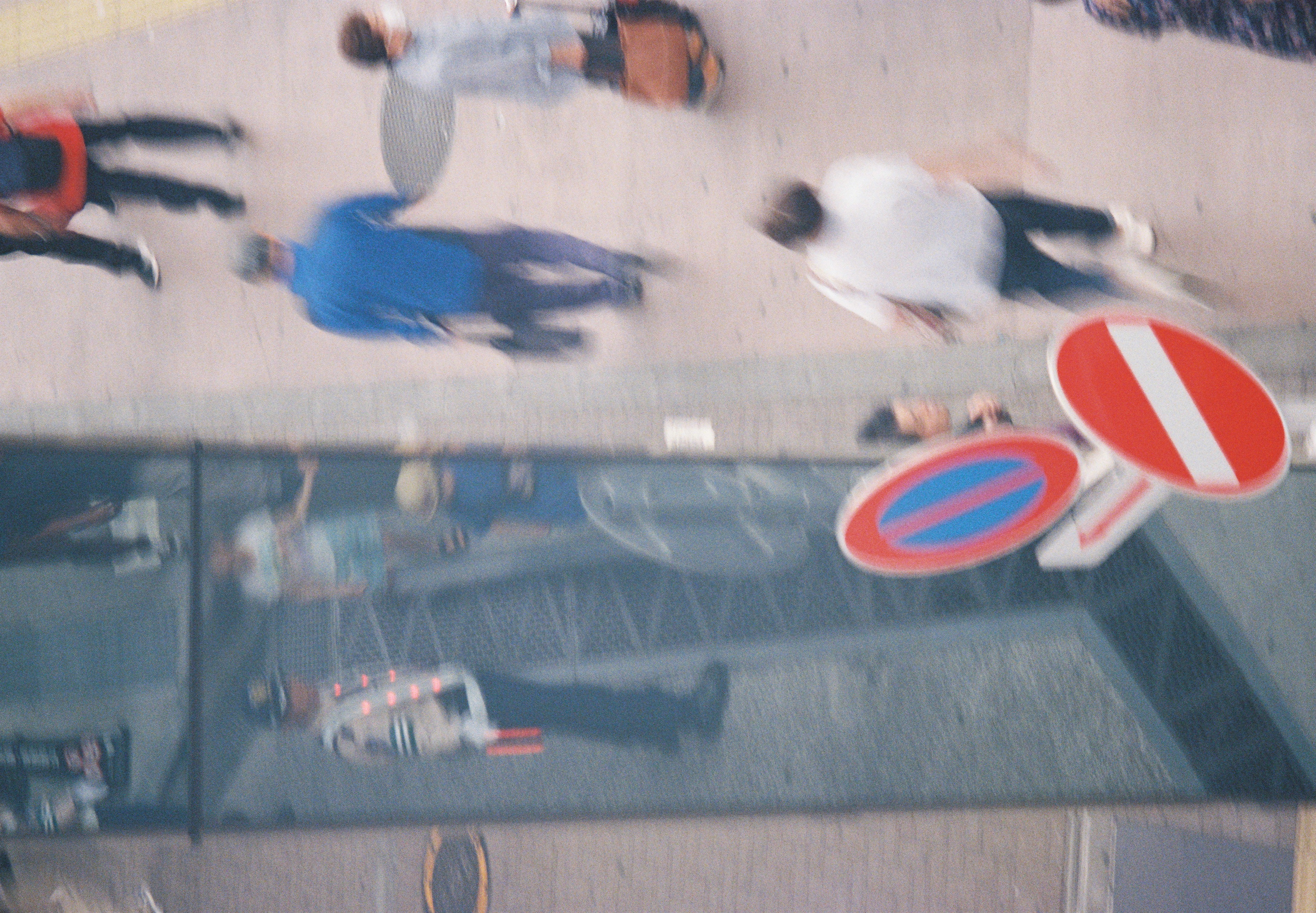 Pedestrians blur past red traffic signs on a sidewalk viewed from above, with a uniformed figure reflected in a glass panel at left.