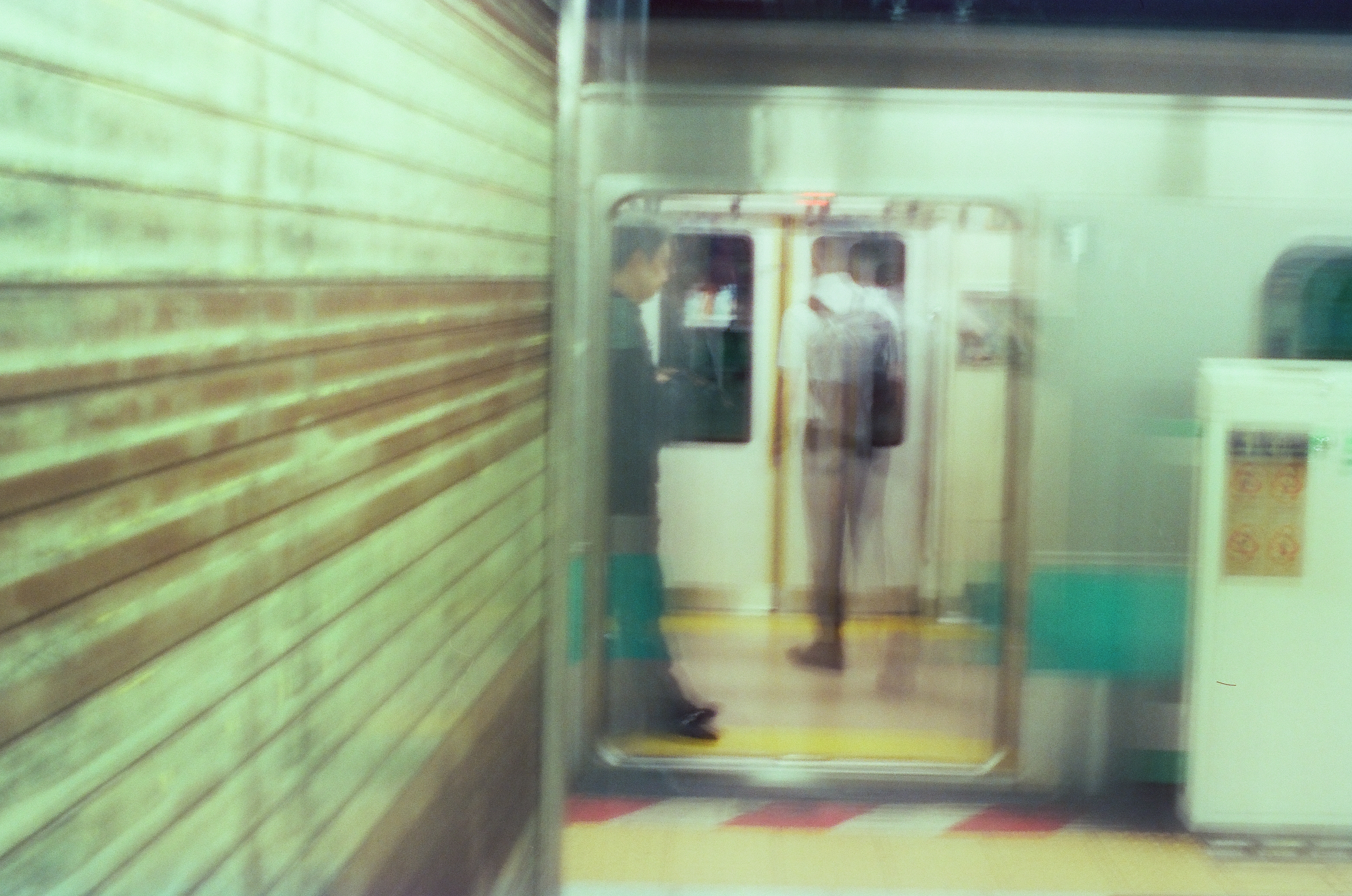 Two passengers stand inside an open train doorway, one in a dark green shirt and the other in a light shirt with a dark backpack, moderately blurred by motion.