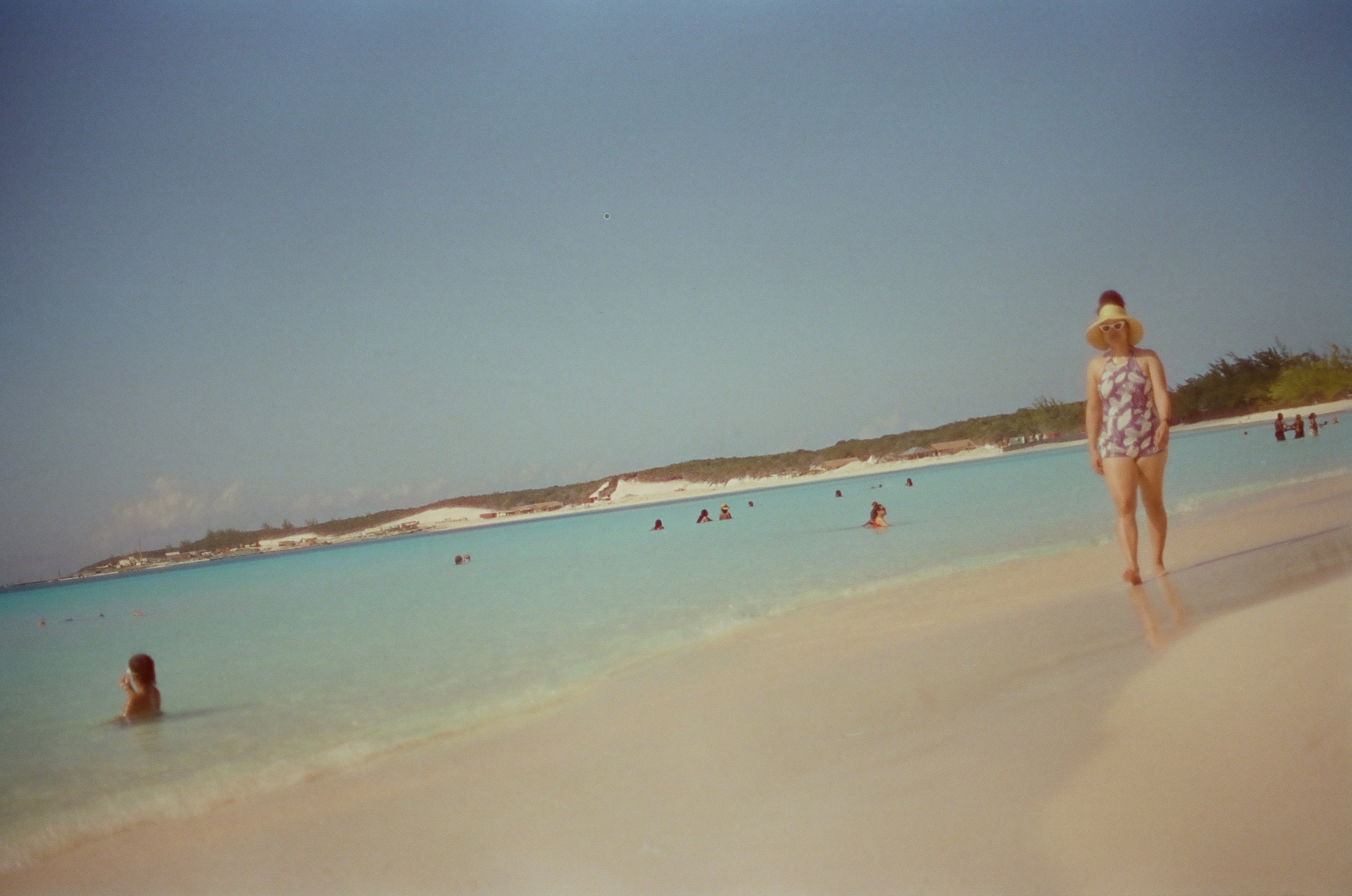 A hat-wearing adult stands beside a tilted shoreline while scattered bathers occupy pale blue water beneath a broad sky.