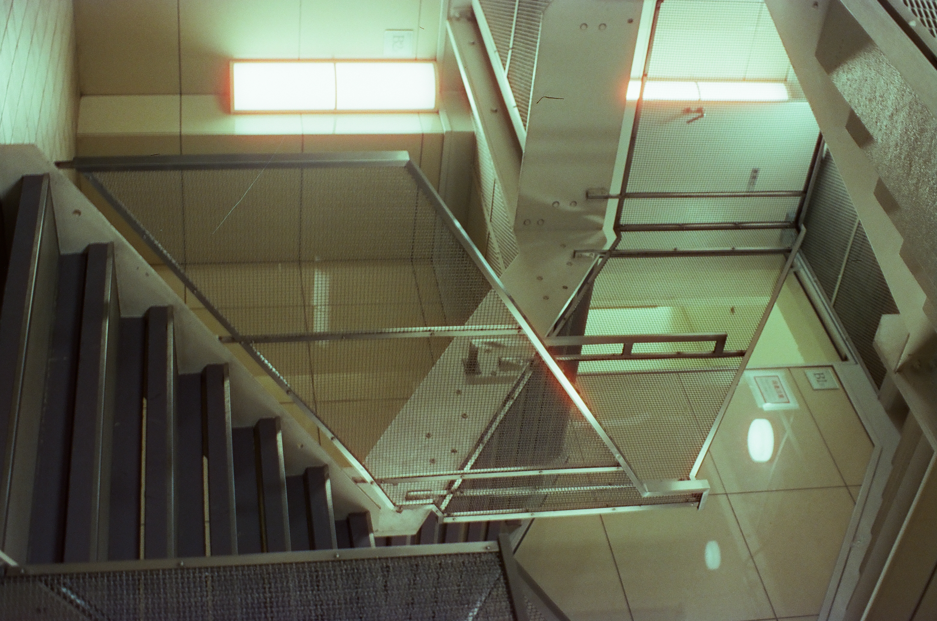 An empty stairwell with gray-edged steps rising to the right, metal handrails, wire-mesh panels, tiled walls, and two glowing rectangular light panels.