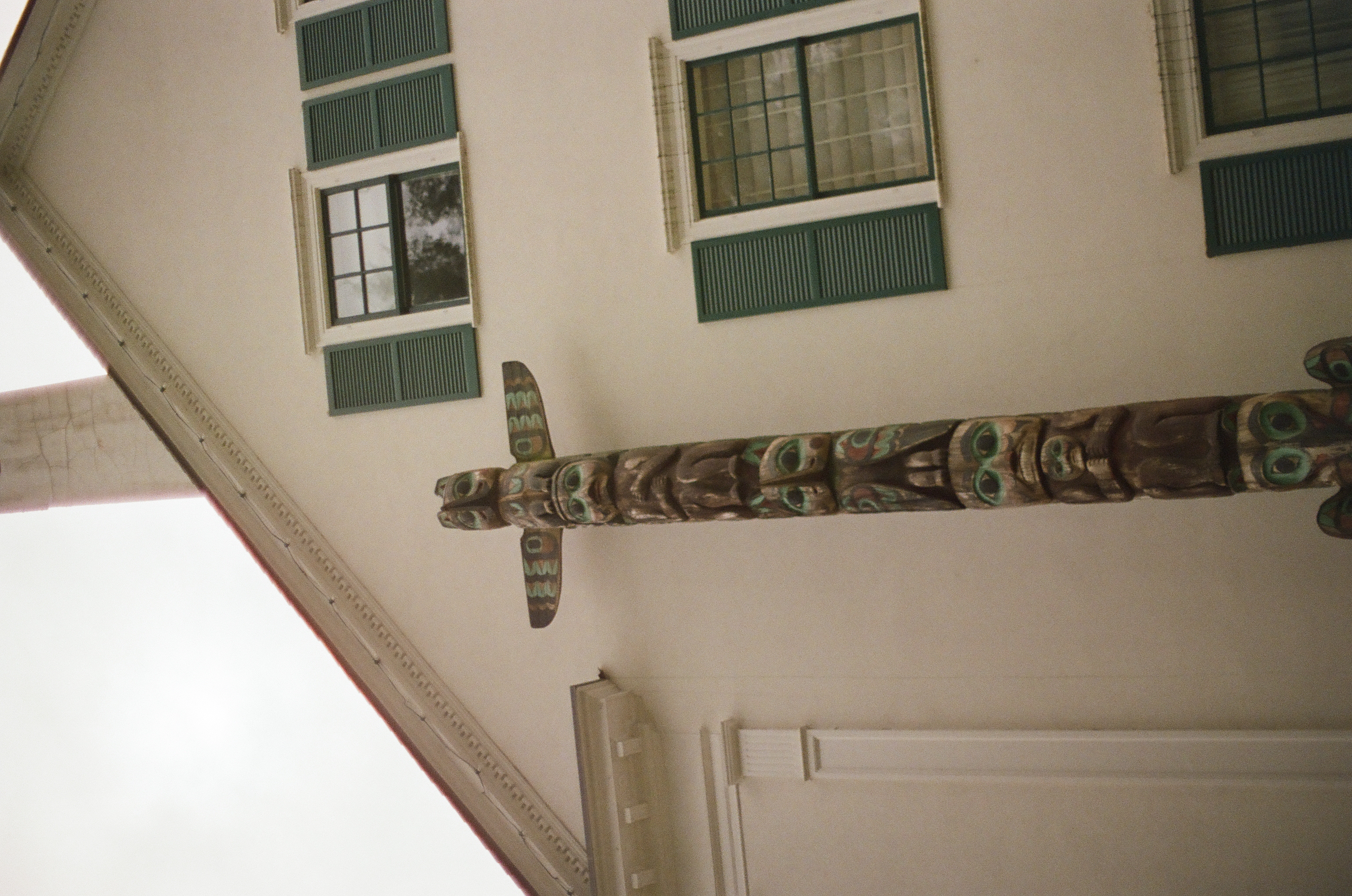 Seen from below, a carved wooden totem with a winged crossbar rises against a cream building with teal-shuttered windows.