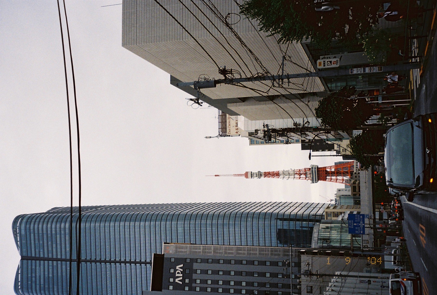 A red-and-white lattice tower rises at the end of a wide avenue between a glass high-rise marked 'APA' and a pale stone building.