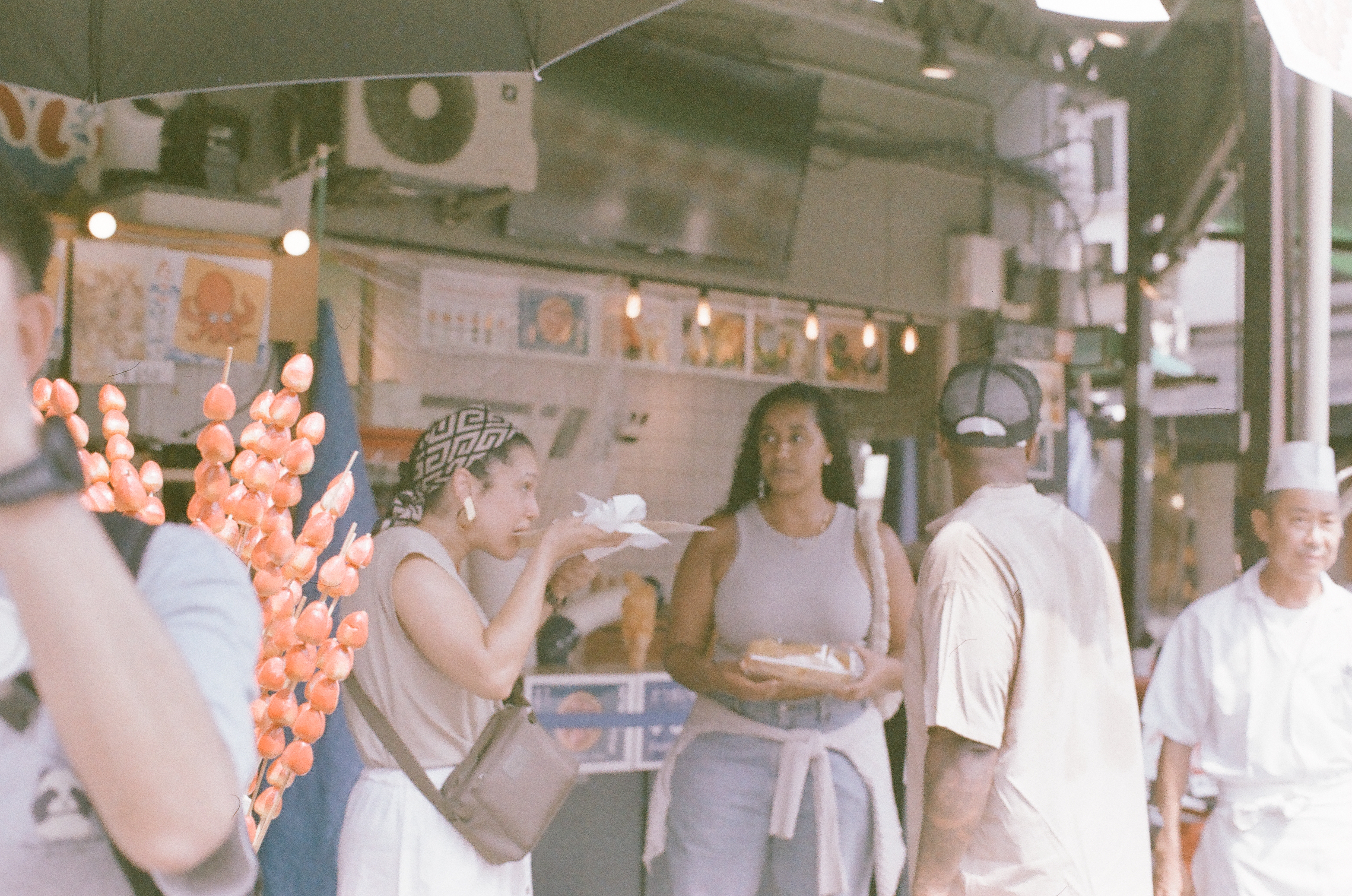 Skewers of glossy orange fruit hang in the foreground as visitors gather near a stall counter under a large umbrella.