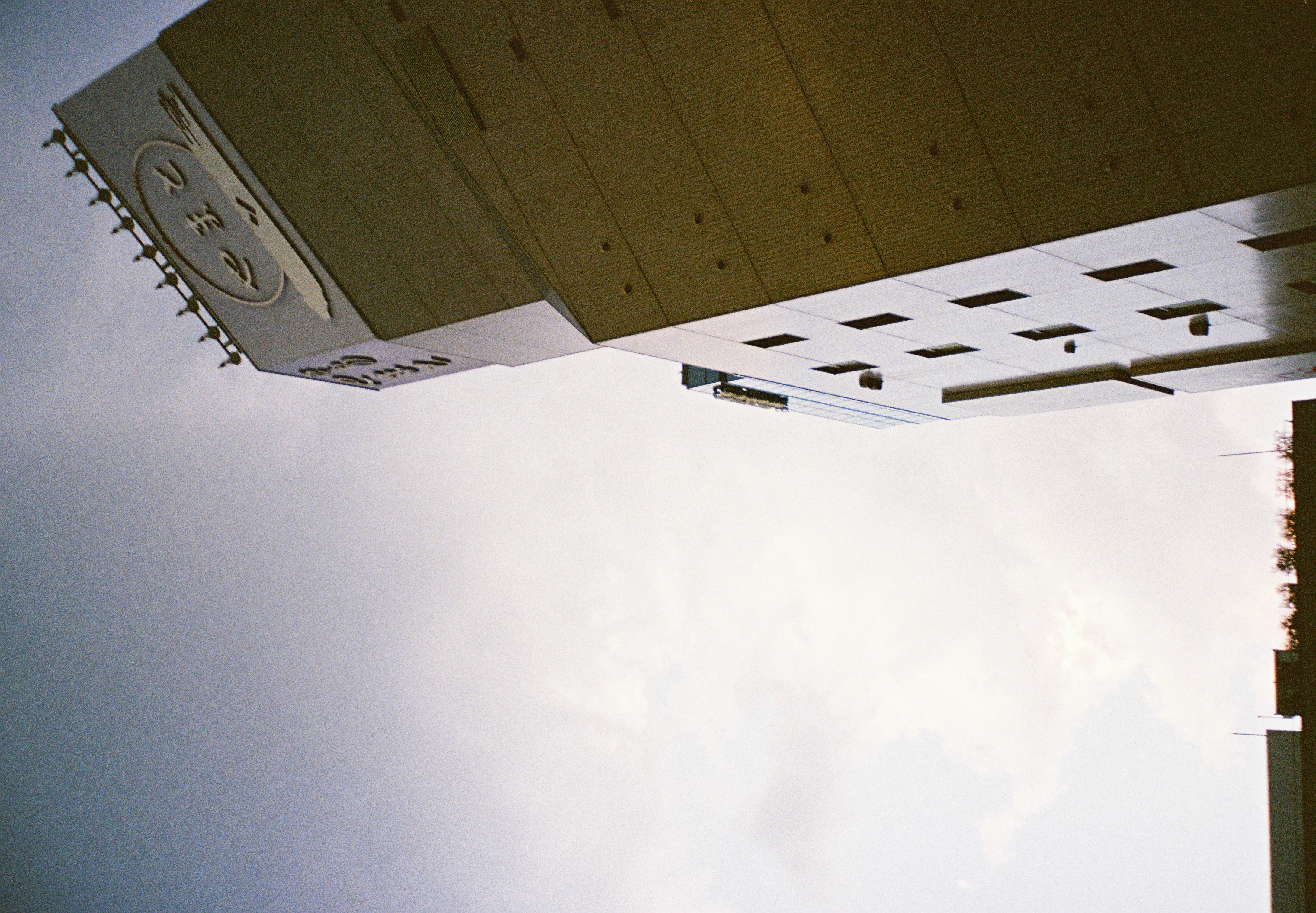 A tall tan-clad building leans across the right side of the frame beneath an overcast sky, topped by an oval sign bearing three Japanese characters.