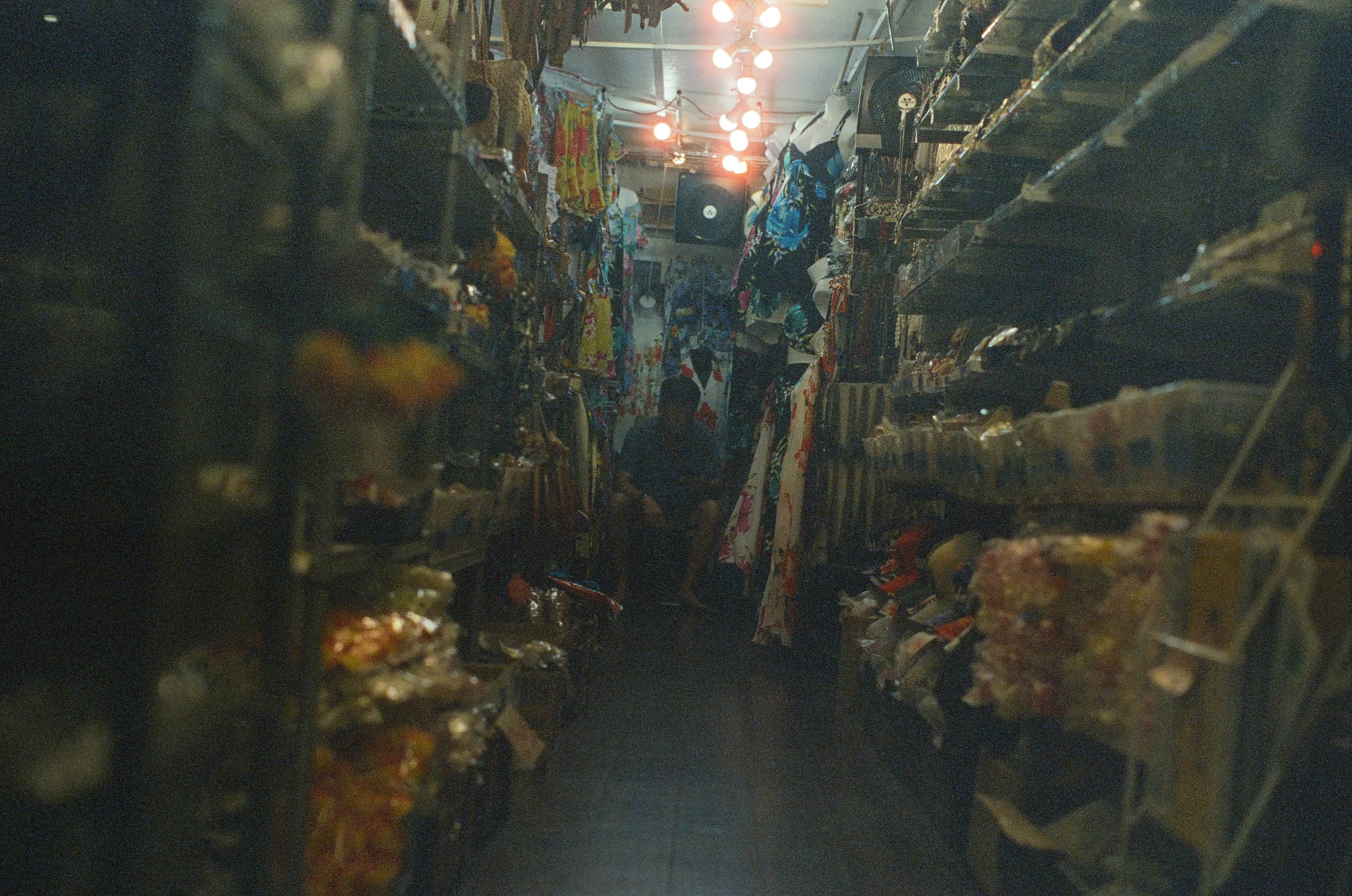 A narrow shop aisle recedes beneath bright bulbs between densely packed hanging and shelved goods.