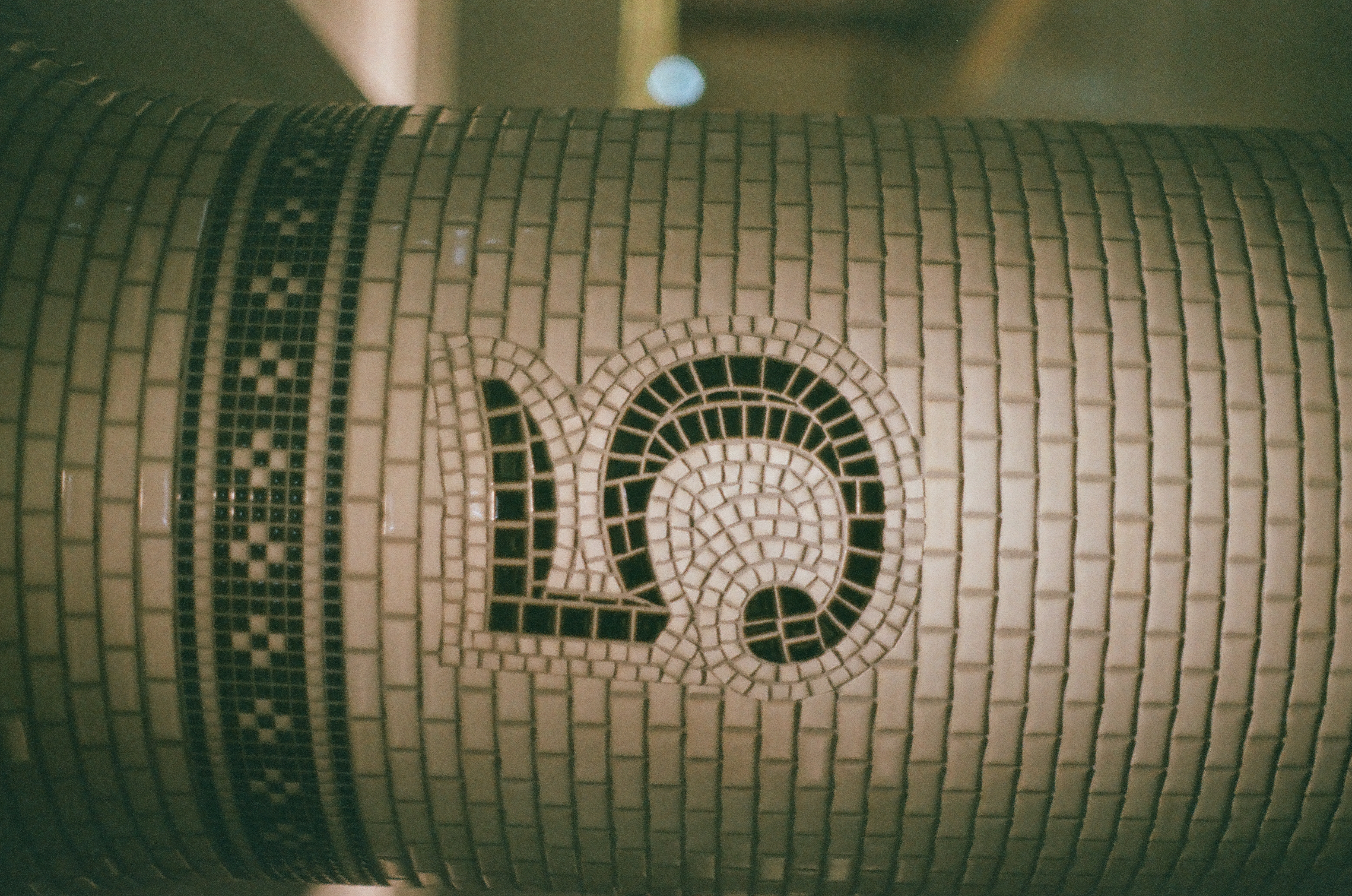 A tiled column displays the numeral five built from dark mosaic tesserae in a flagged, curling shape beneath a checkered band, with a small blue light blurred nearby.