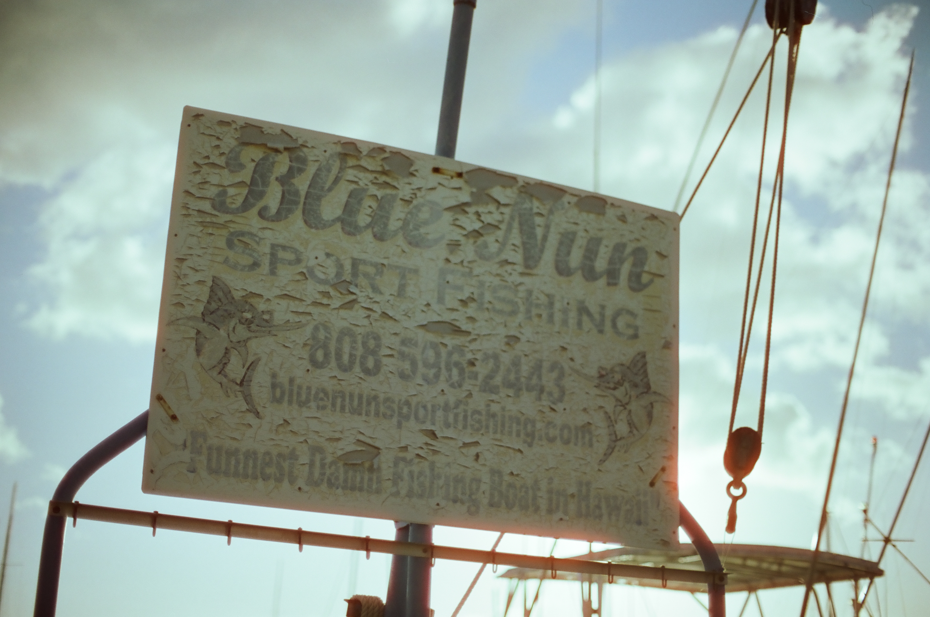 A weathered fishing-charter sign hangs between ropes and rigging beneath a bright blue and white sky.