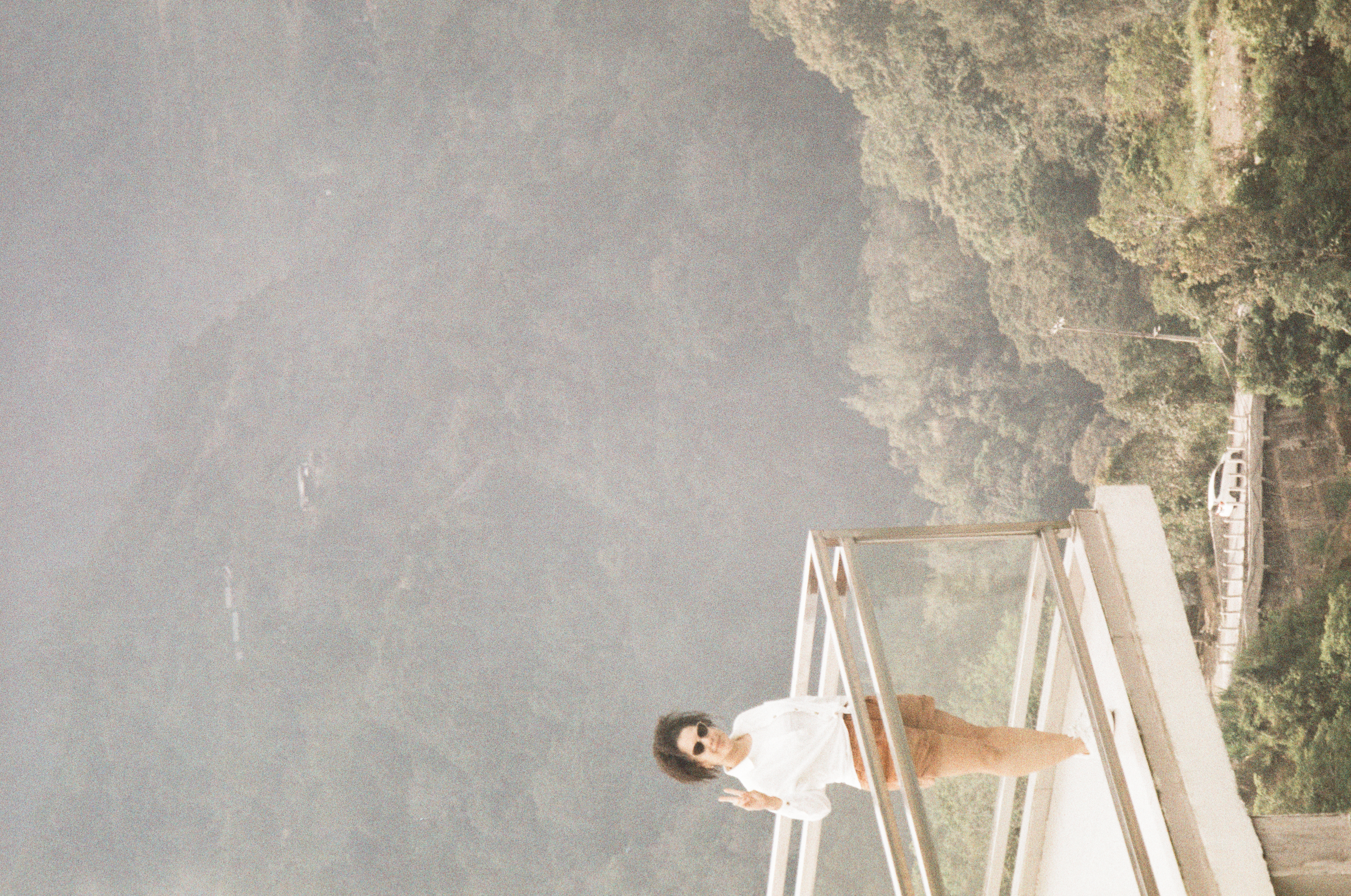 A person in sunglasses, a white top, and brown shorts raises two fingers beside a glass railing above wooded slopes.