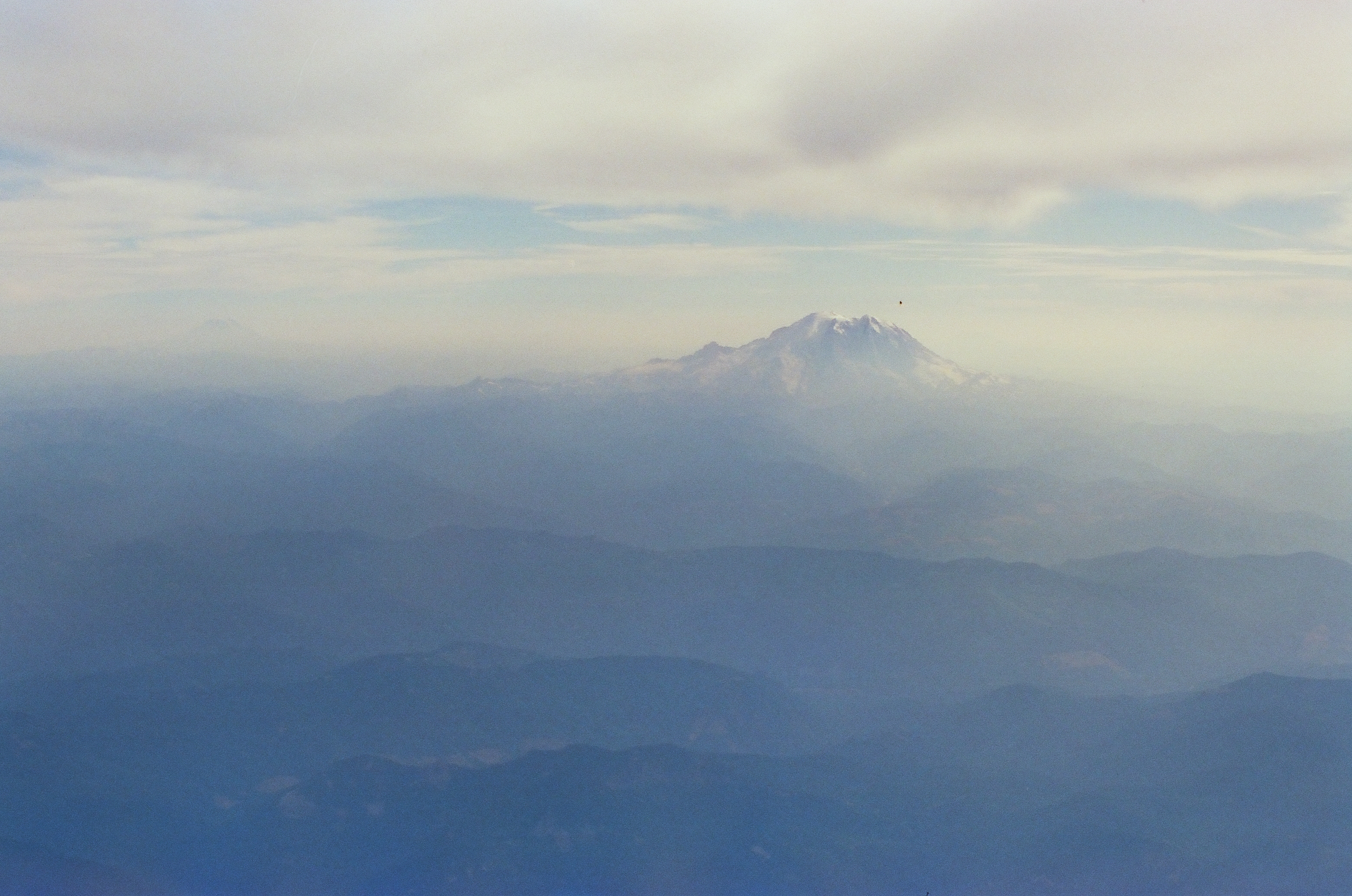 A snow-capped peak partly wrapped in cloud rises above layered hazy ridgelines, with a small dark mark visible in the sky near its top.