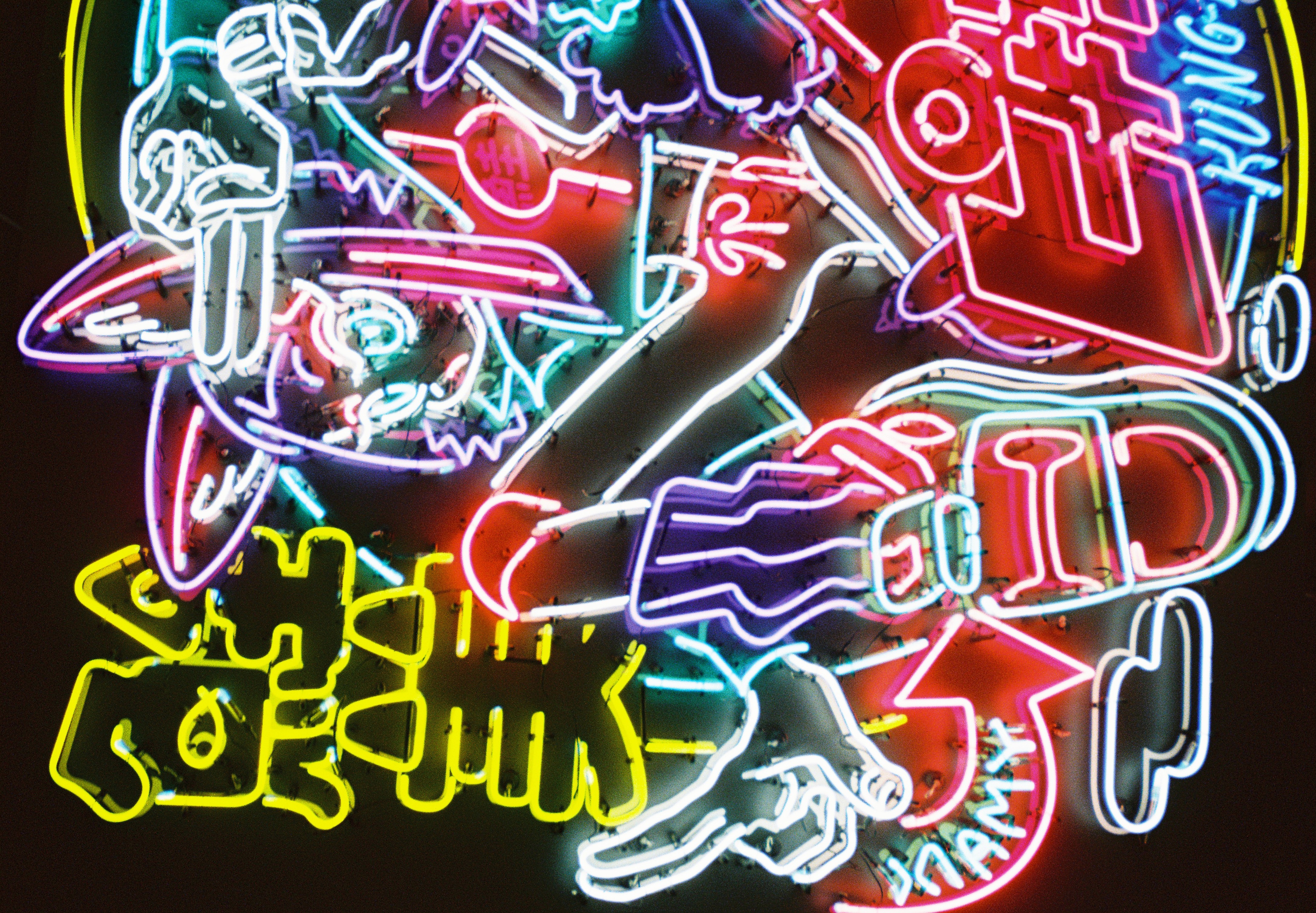 Close view of a neon sign showing a cartoon animal character with pointed ears, two raised fingers, and chunky sneakers in pink, white, teal, and yellow tubing.