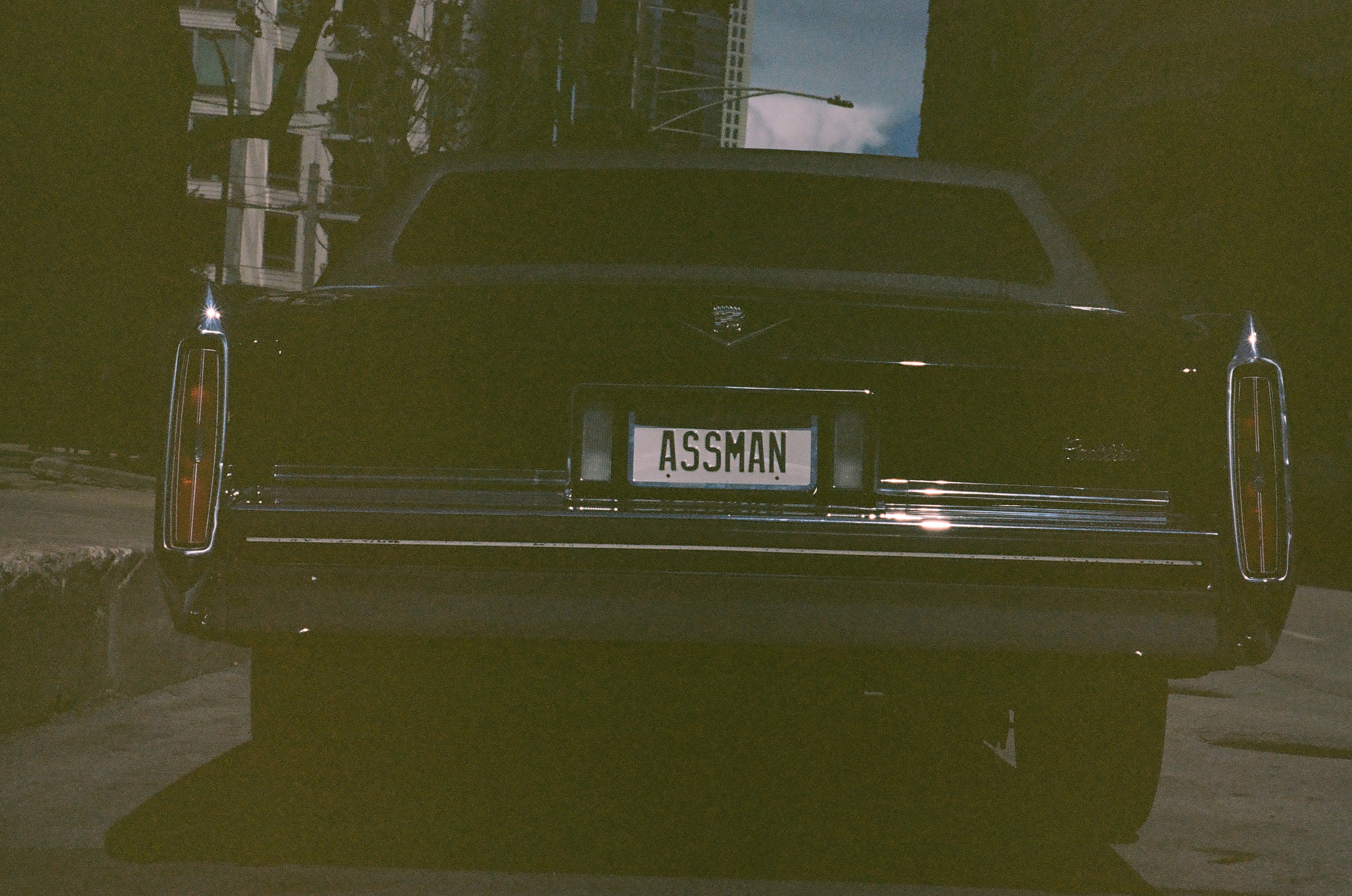 The polished rear of a black vintage car fills a narrow curbside view between pale pavement and buildings.