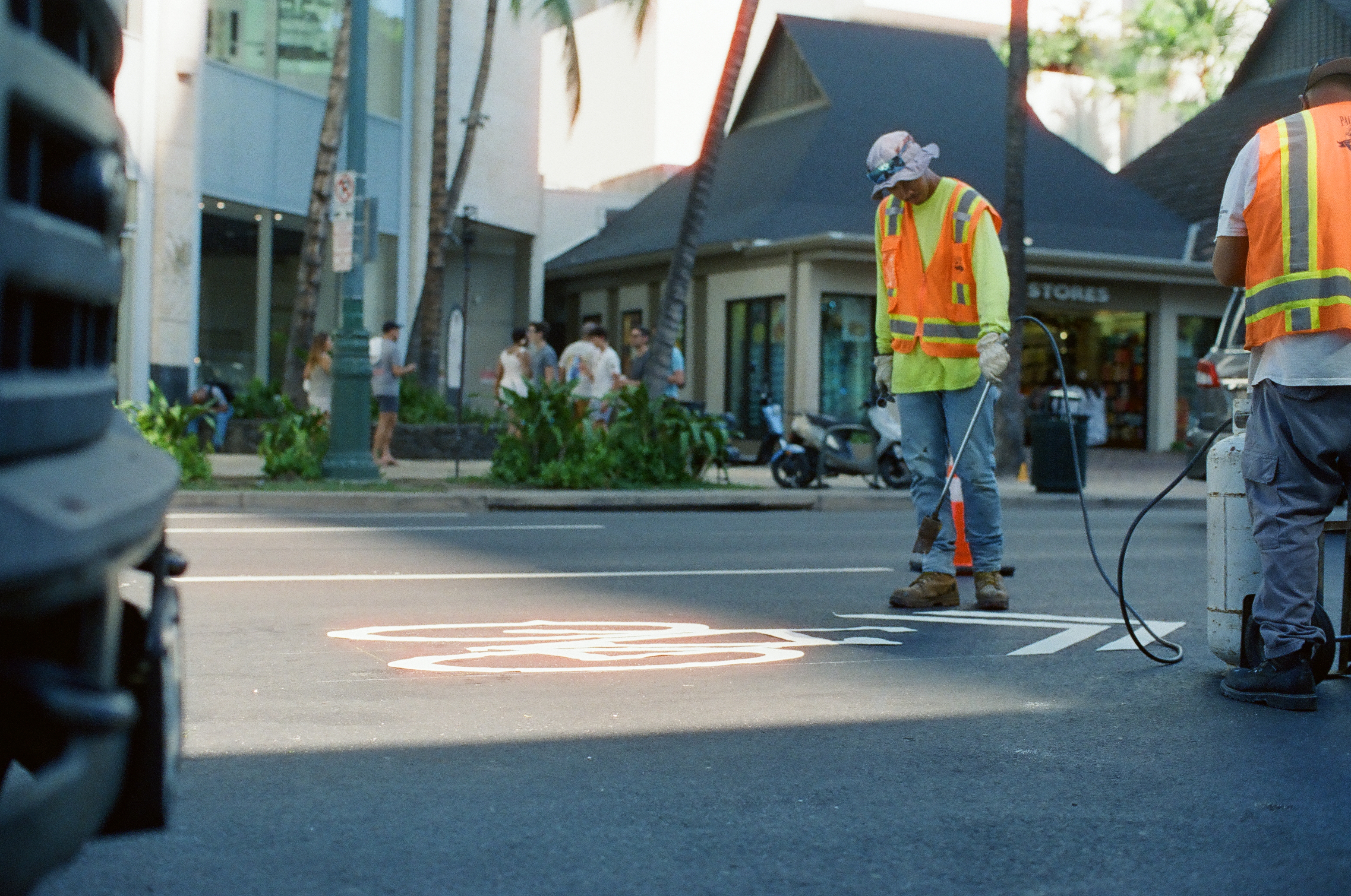 A worker in an orange vest and yellow sleeves paints a white bicycle symbol on the asphalt while a colleague operates a hose-fed machine nearby.