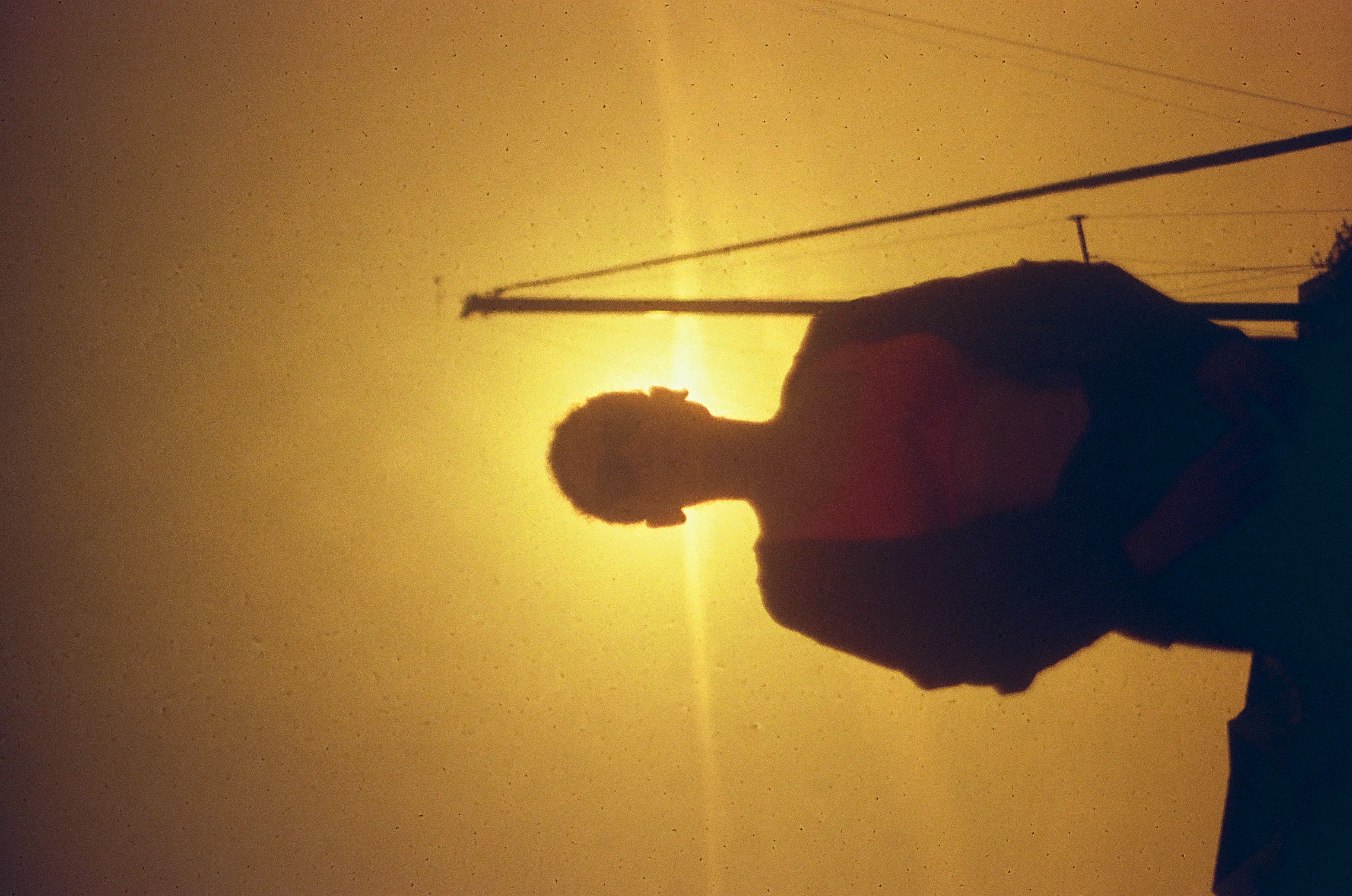 A person's silhouetted head and shoulders stand before a blazing amber sun, with utility wires and a pole crossing the glowing sky.