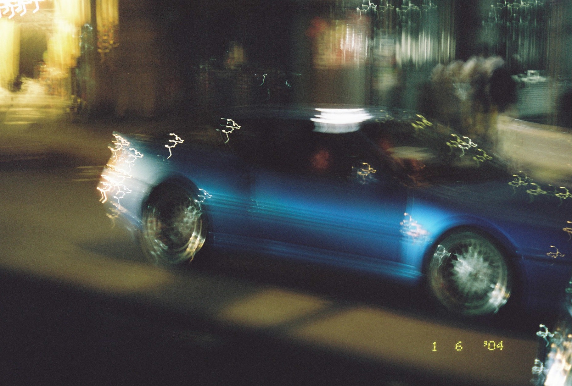 A deep blue car crosses a night street, motion blur stretching its wheels and brightwork into loops of light against a glowing background.