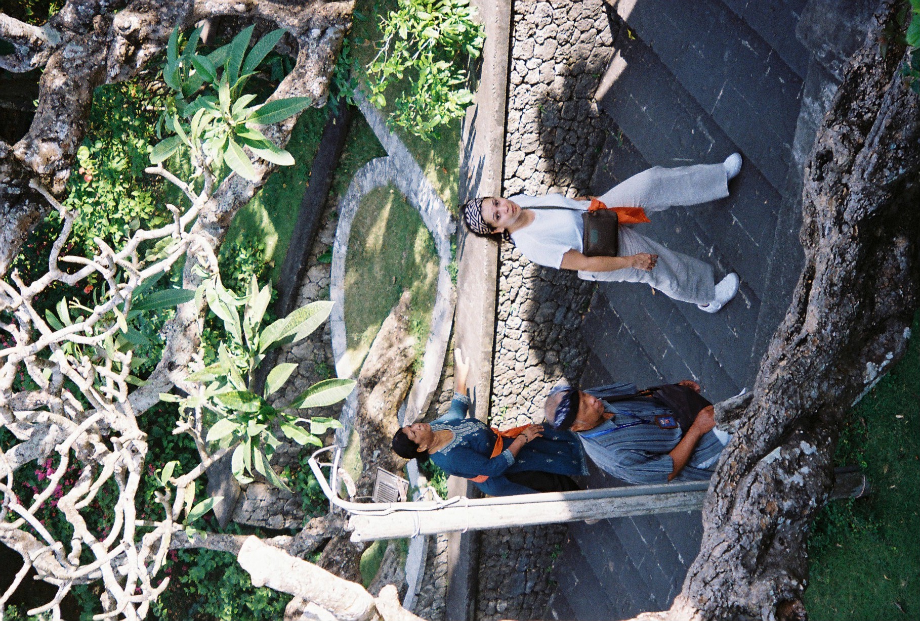 Three people in traditional dress and head cloths stand beside a circular stone ring set into a grass lawn, shaded by frangipani branches.
