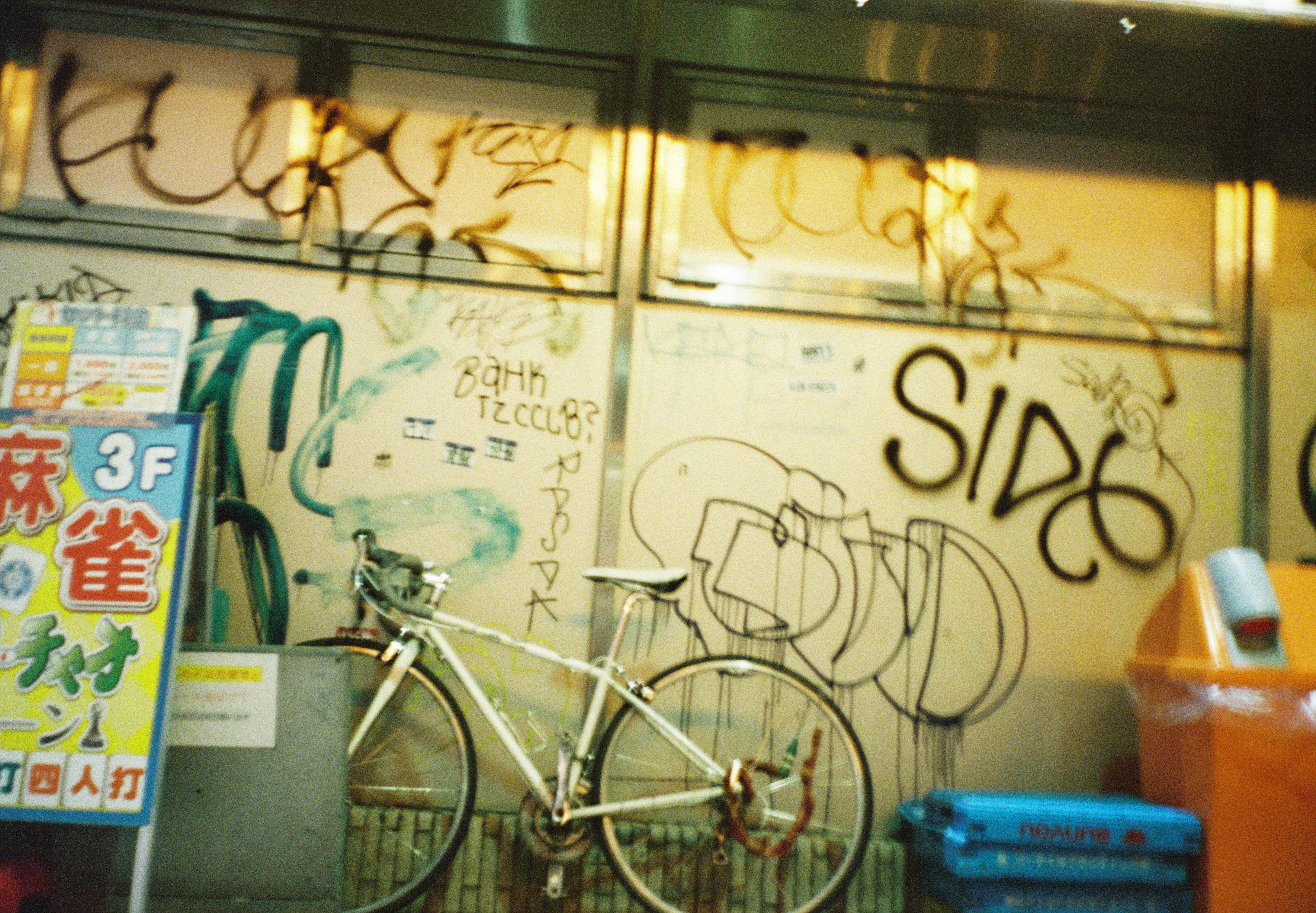 A white road bicycle leans against a pale wall covered in black and green spray-painted tags, with blue crates and an orange bin at right.