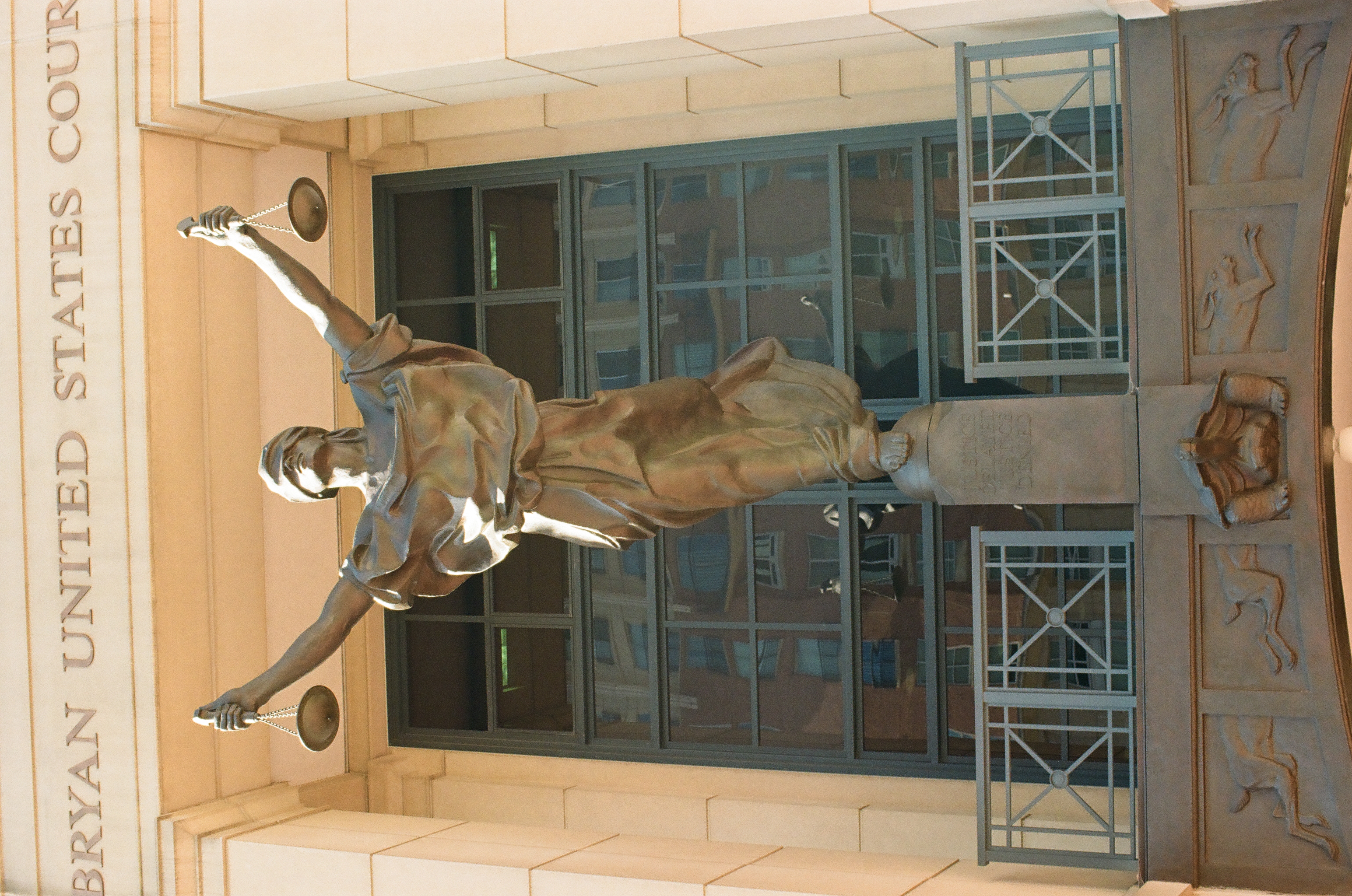 A blindfolded bronze figure holds raised scales in each hand while standing on a pedestal in front of a courthouse's gridded glass window.