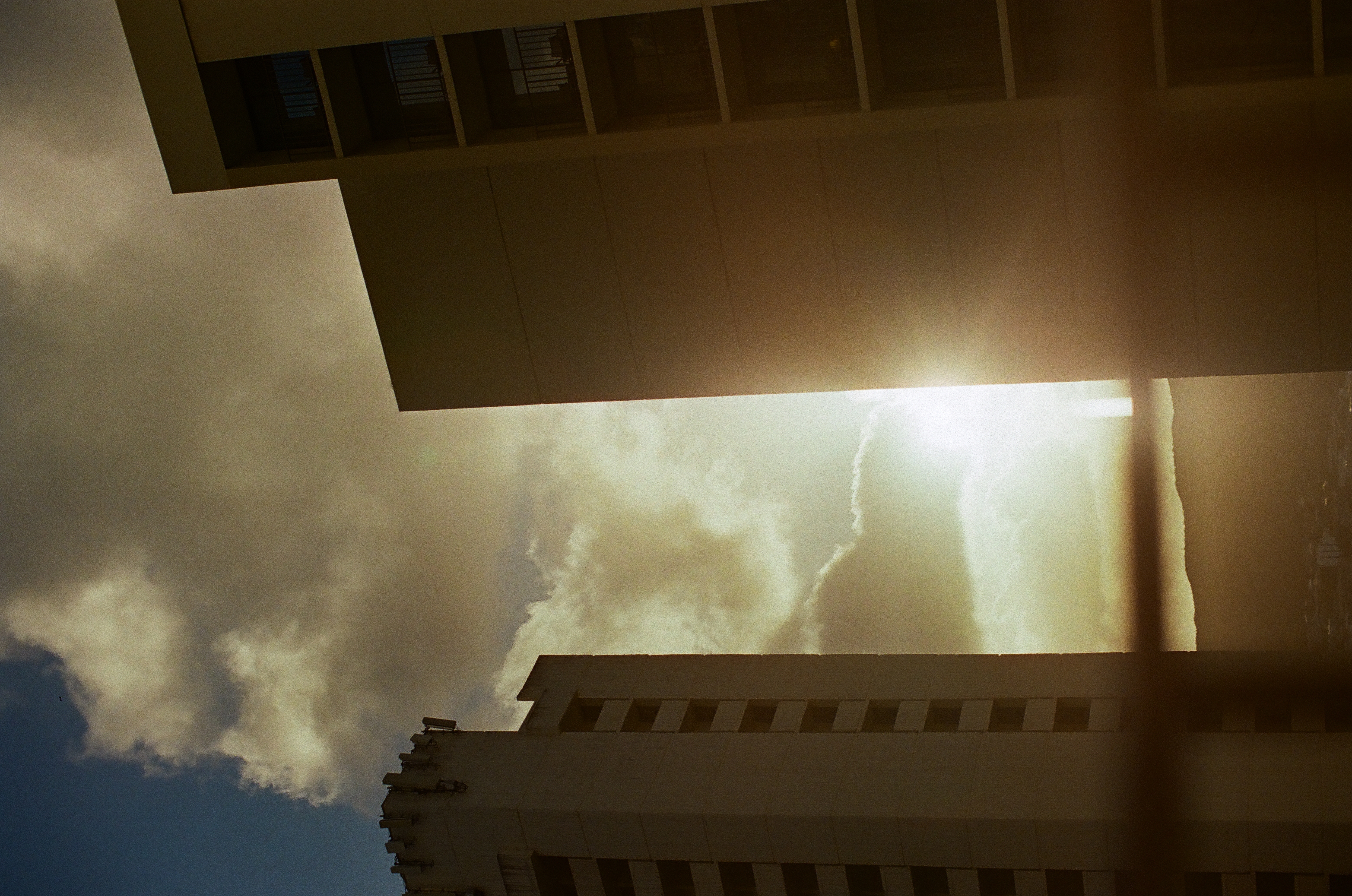 Bright sun breaks between tall dark buildings beneath large white clouds and a narrow blue sky.