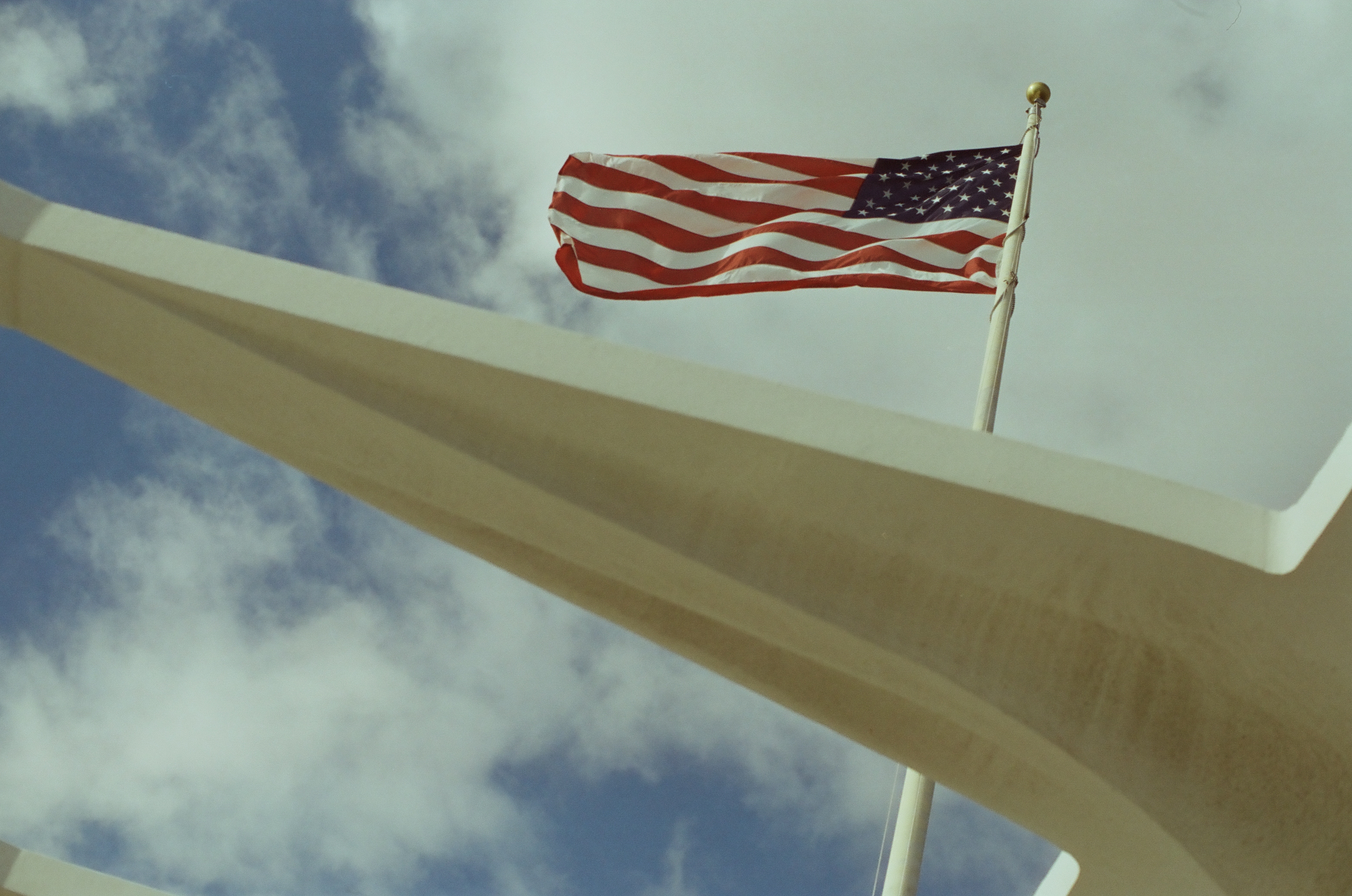 An American flag waves above a sweeping white concrete arch against a bright clouded blue sky.